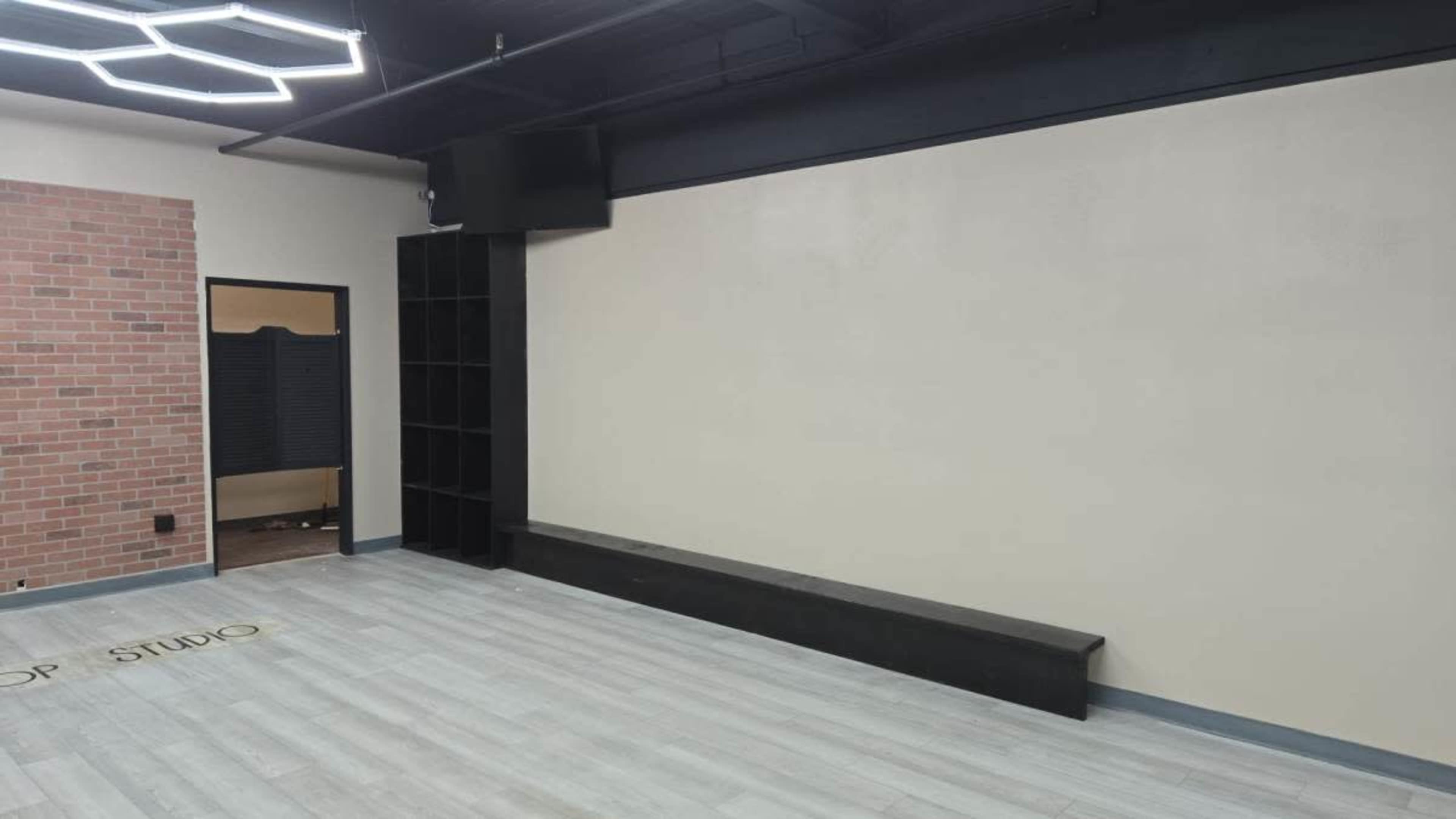 The image shows an empty interior space with a light-colored wall, a black shelf unit, and a grey wooden floor.