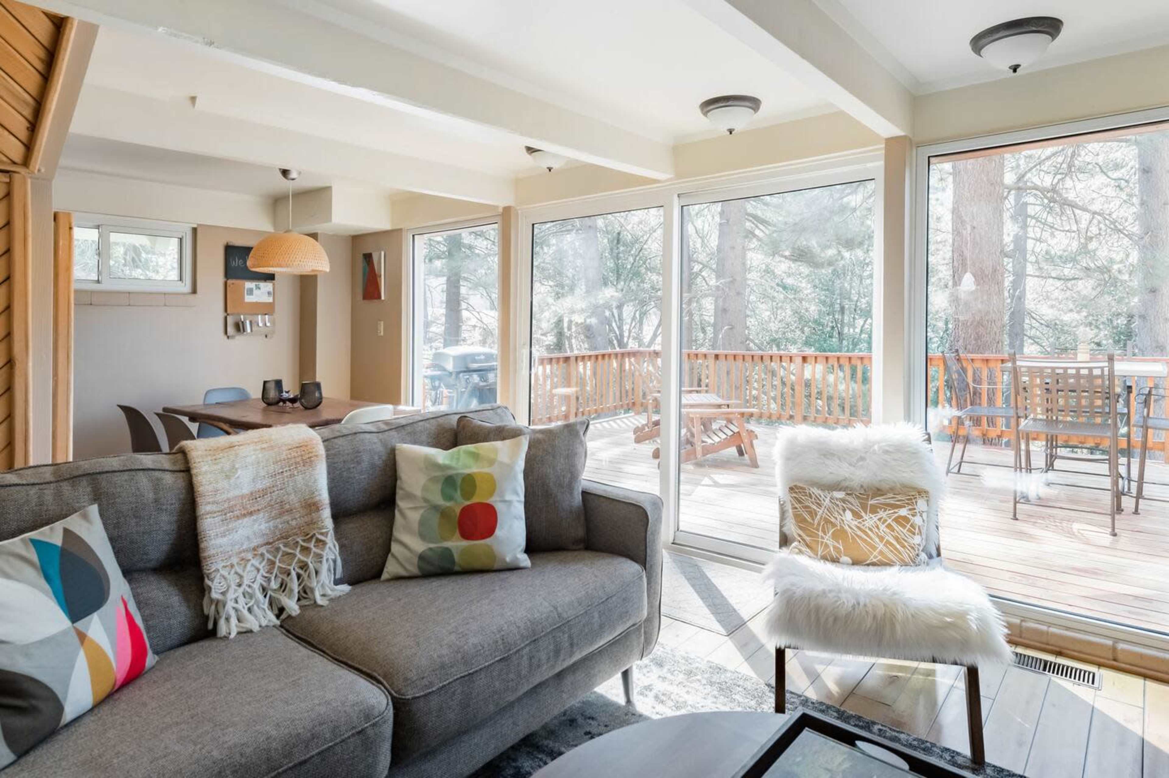 A living room with large windows overlooking a wooden deck and trees, featuring a gray sofa with colorful pillows and a round coffee table.