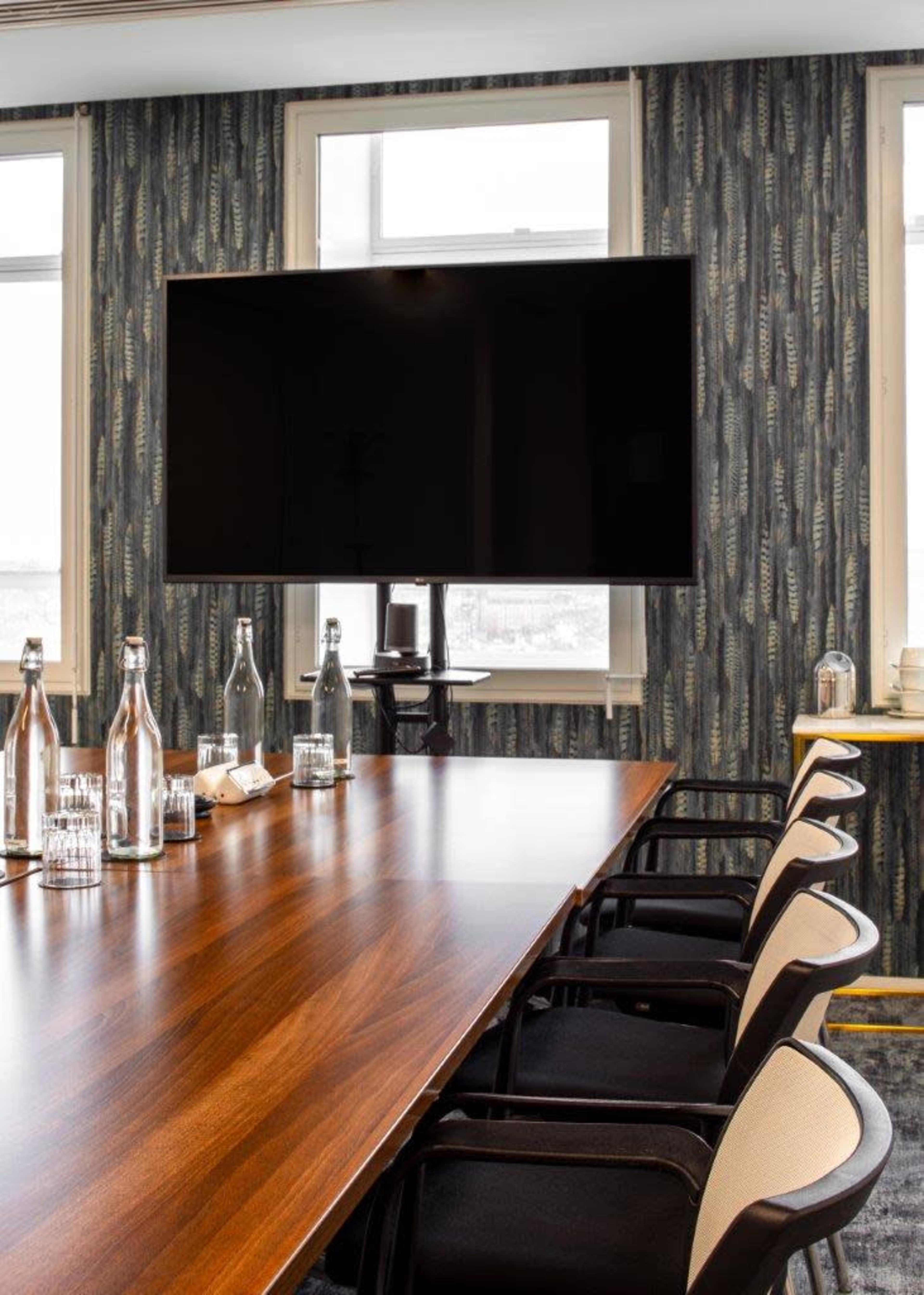 A modern conference room features a long wooden table, black chairs, a large mounted television, and windows with patterned wallpaper.