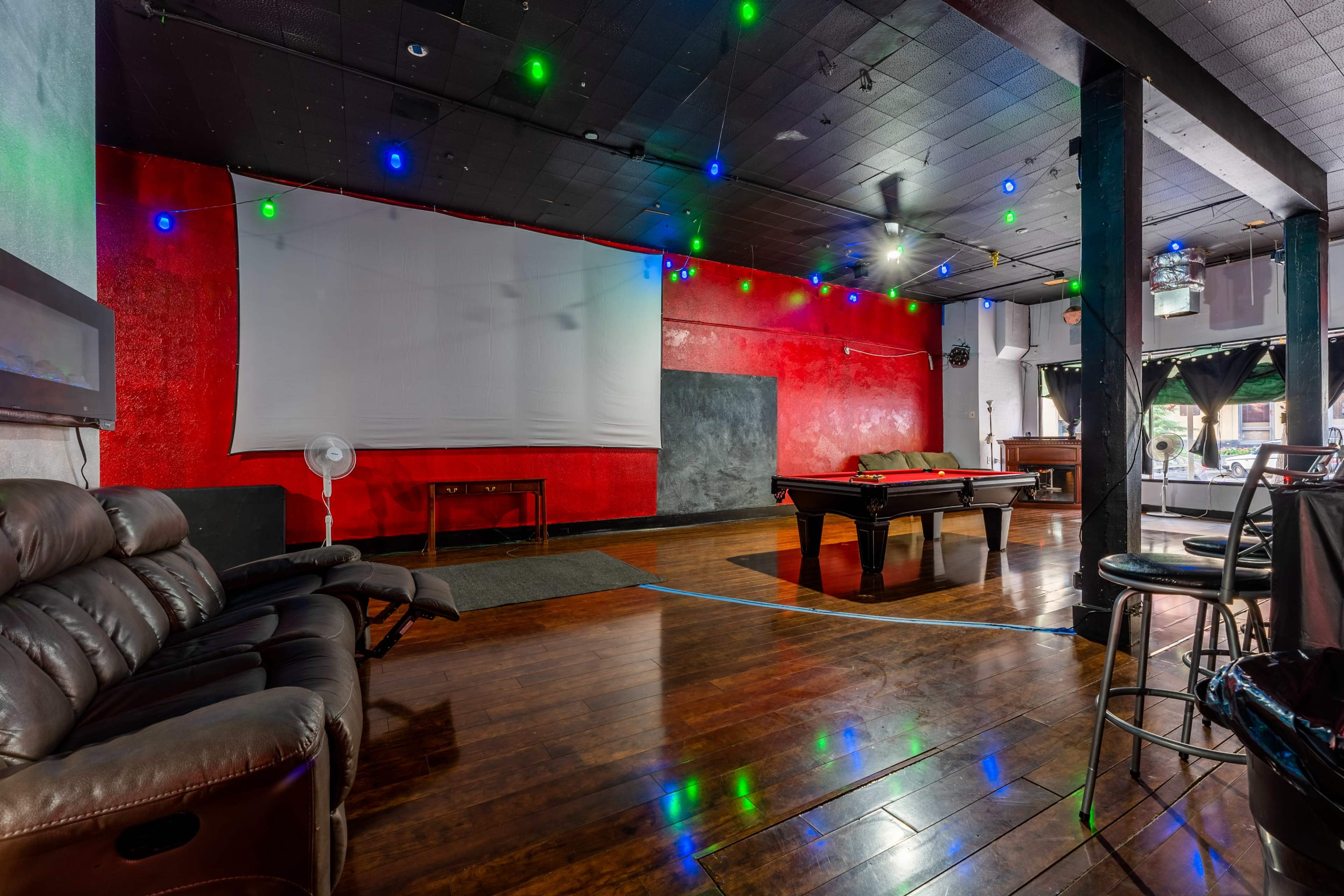 A spacious recreational room with a pool table, a large screen on the wall, and colorful lights strung across the ceiling.