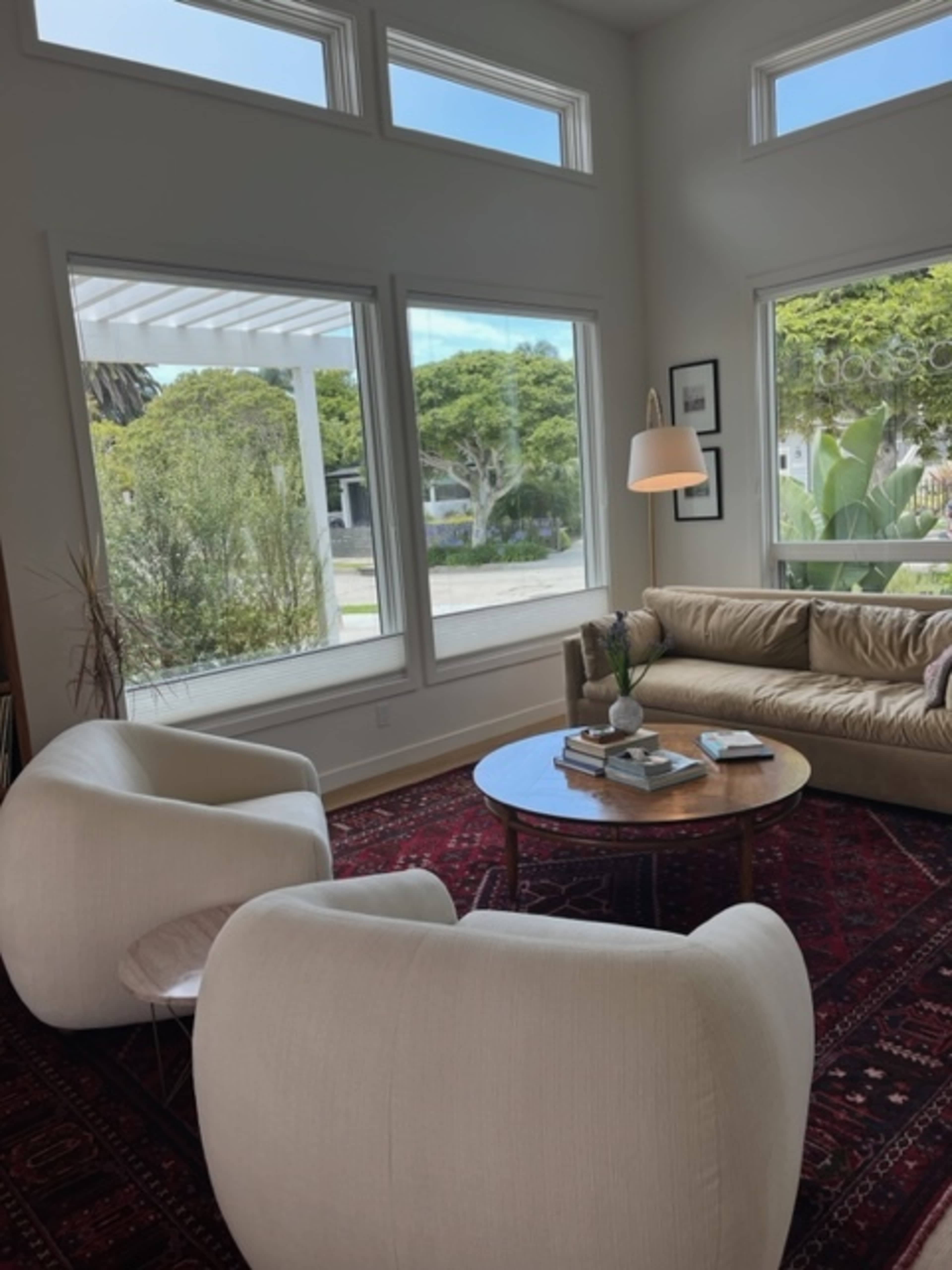 A bright living room with two cream-colored chairs, a beige couch, and a round coffee table, all arranged near large windows that overlook a green landscape.