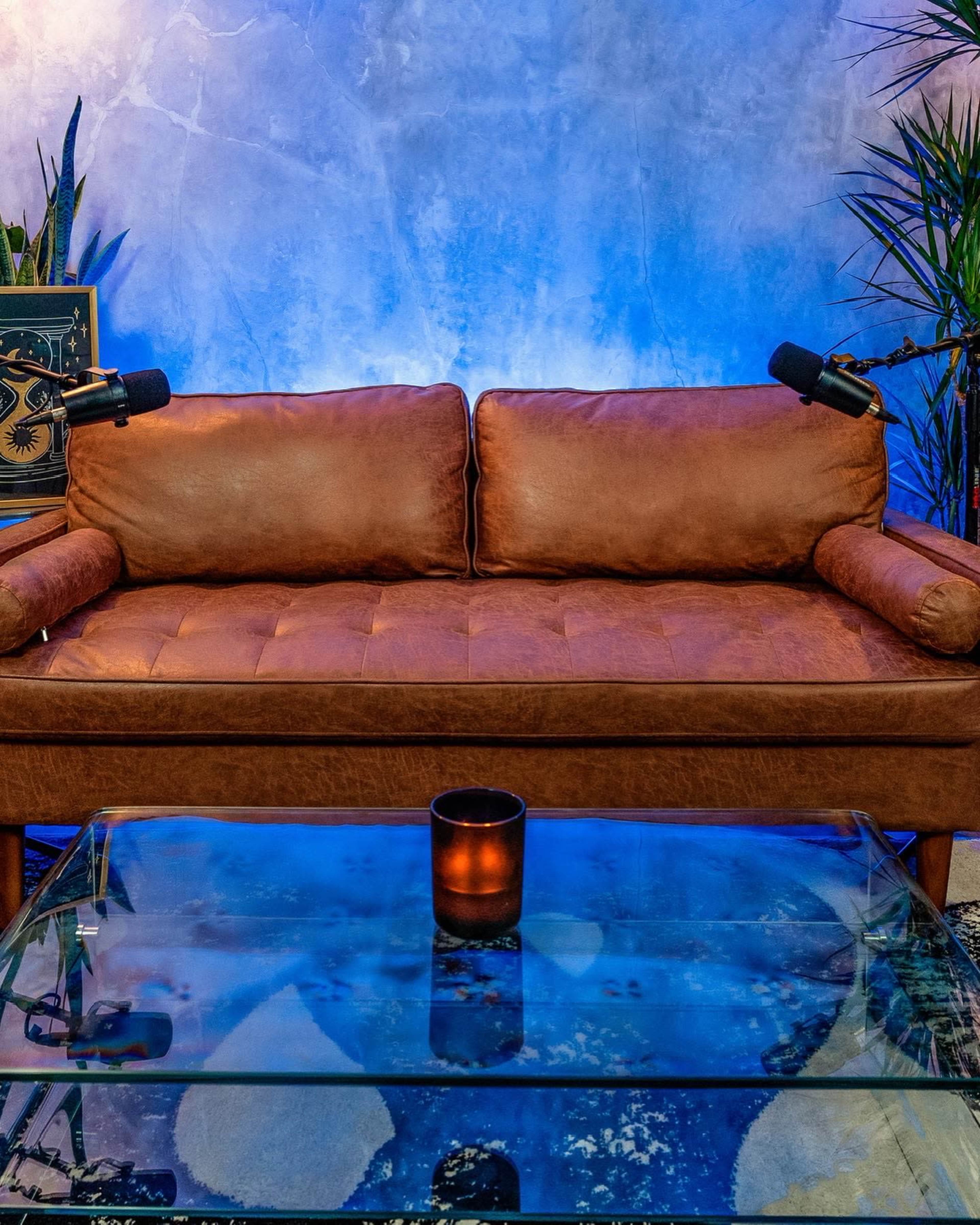 A brown leather sofa is positioned in front of a glass coffee table, with two microphones mounted on the sides and blue lighting illuminating the background.