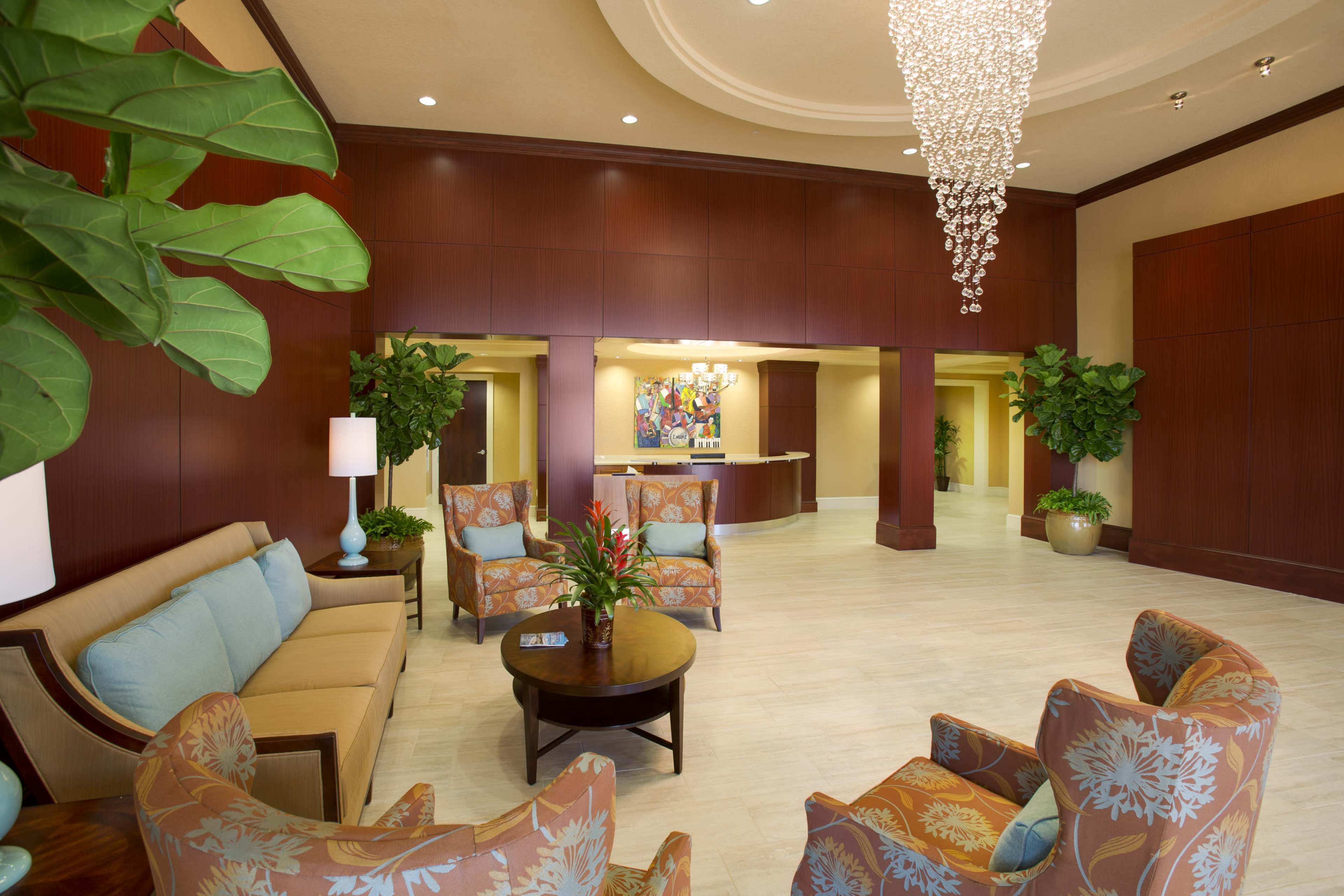 The image shows a brightly lit lobby area featuring a seating arrangement with patterned chairs, a coffee table, and decorative plants, alongside a reception desk in the background.