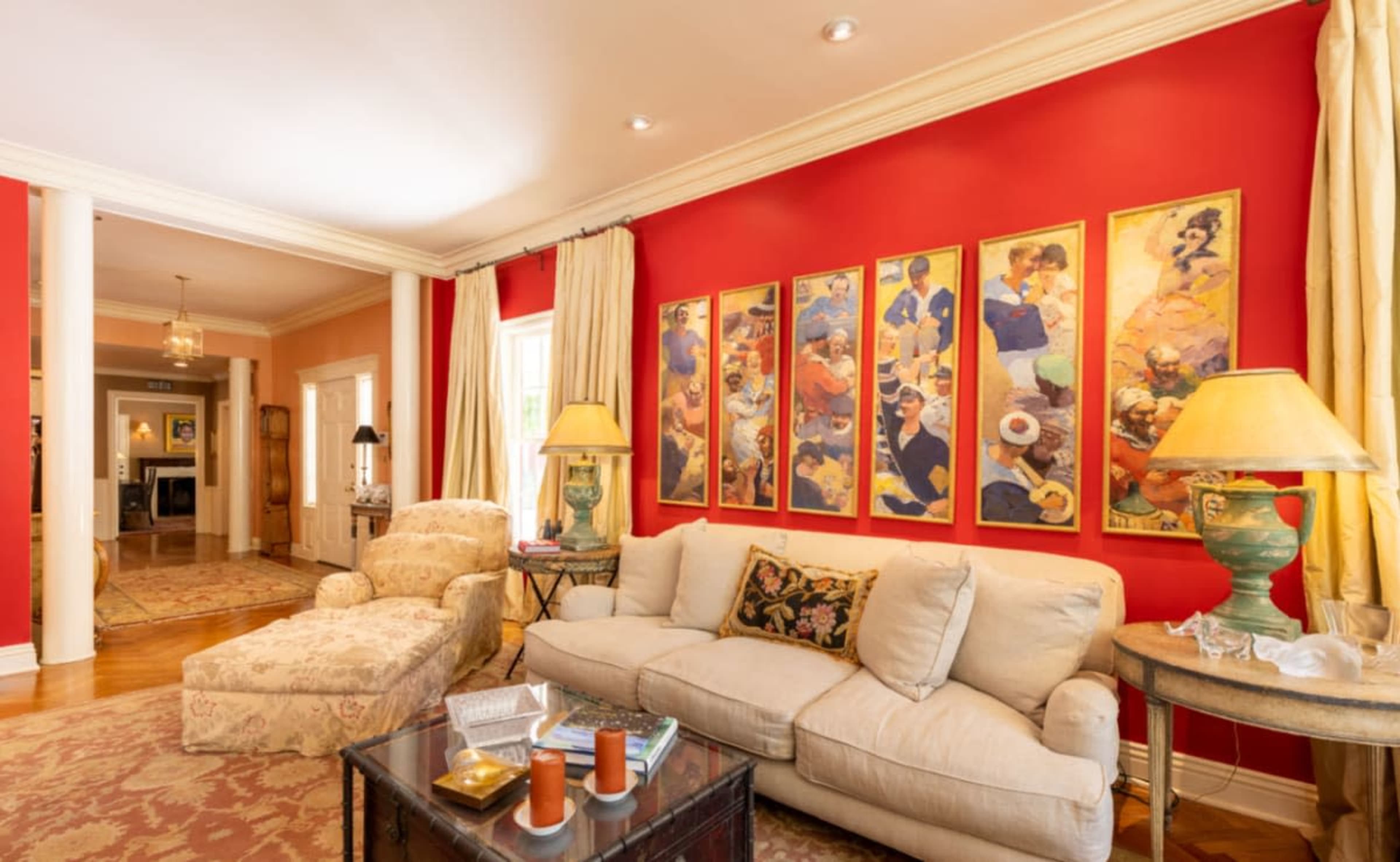 A living room with a beige sofa and a patterned chaise lounge against a red wall adorned with colorful artwork.