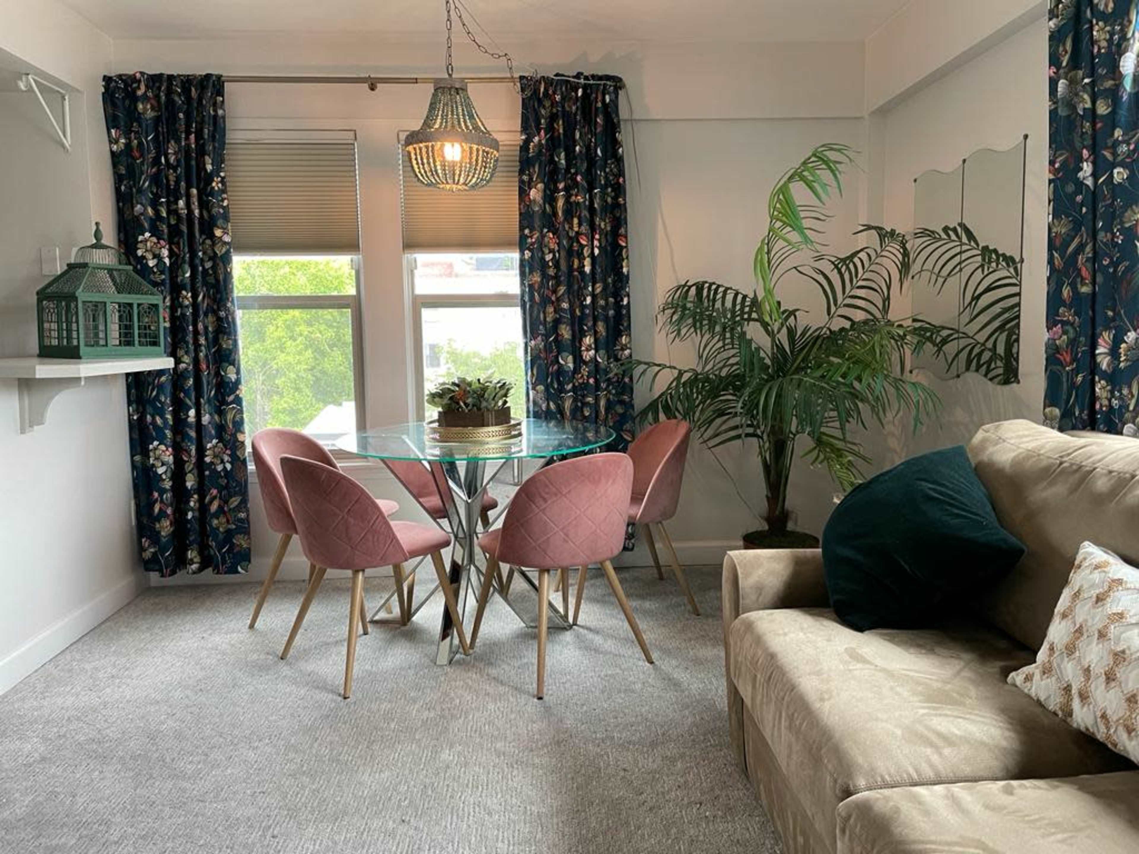 The image shows a living area with a beige sofa, a glass dining table surrounded by pink chairs, and large windows covered with floral curtains.