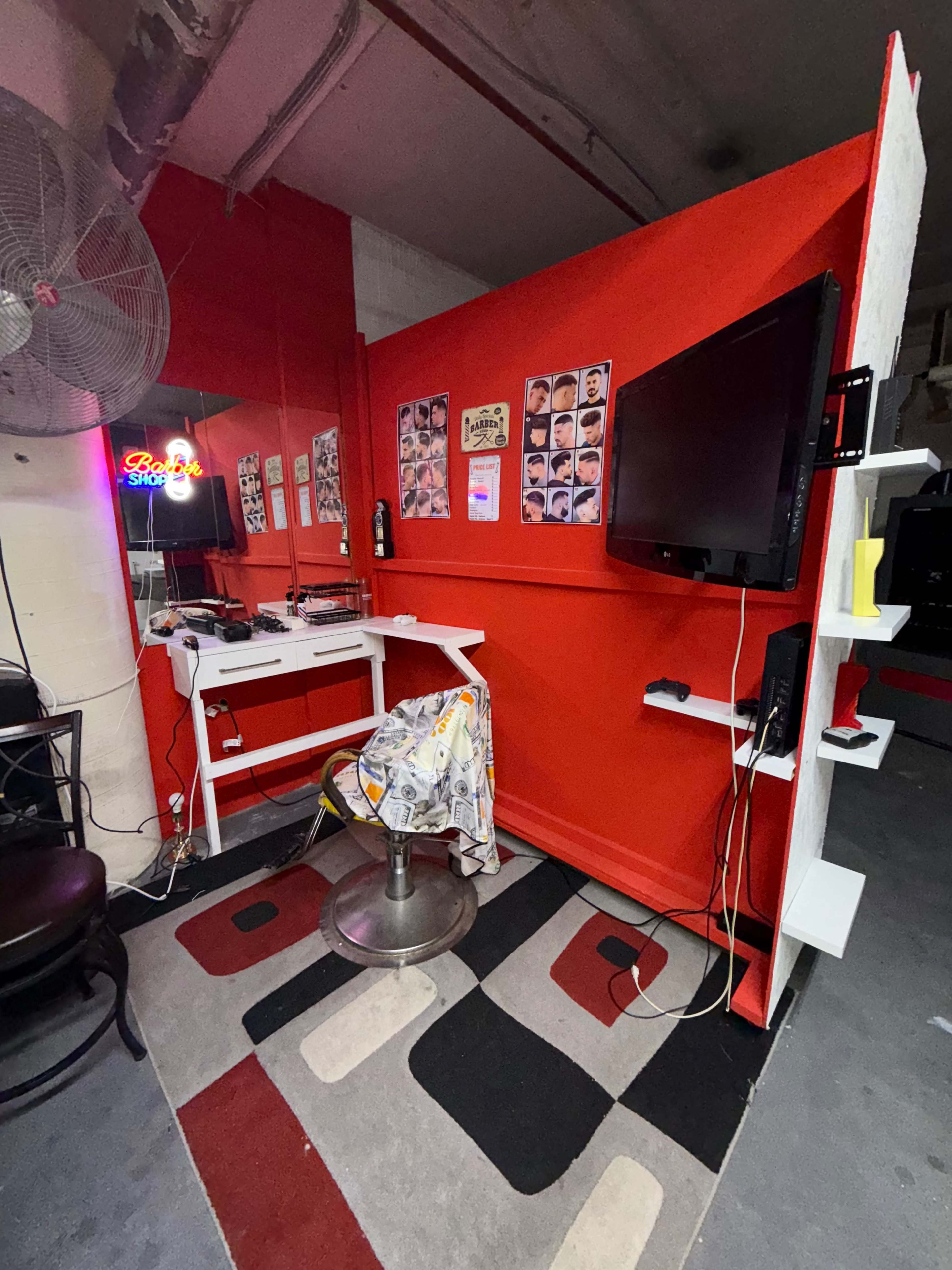 The image shows a small haircutting area with a red wall, a black TV mounted on the side, a circular stool, and various photographs on the wall.