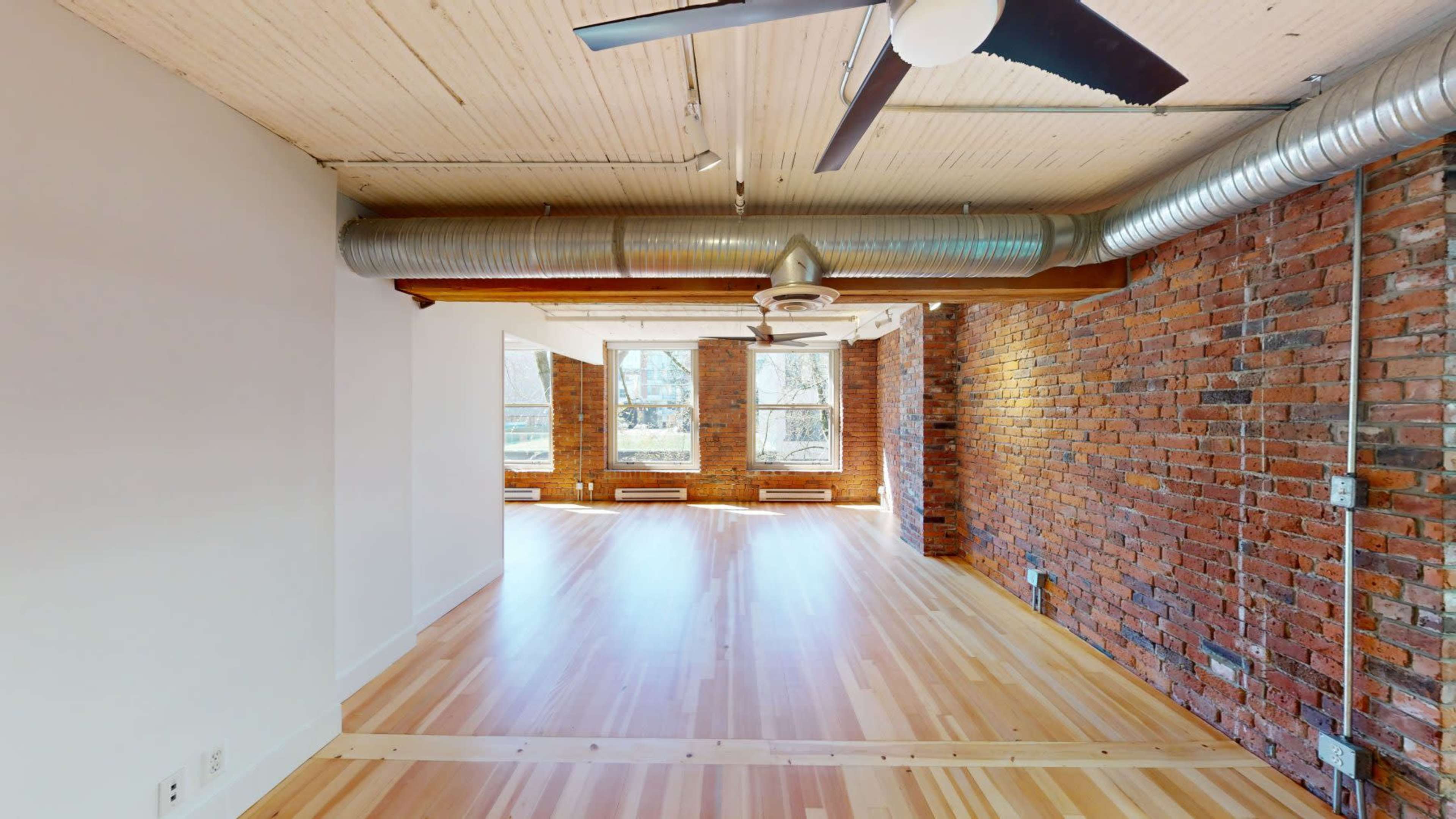 The image shows a spacious interior of a modern loft with exposed brick walls, wooden flooring, and large windows.