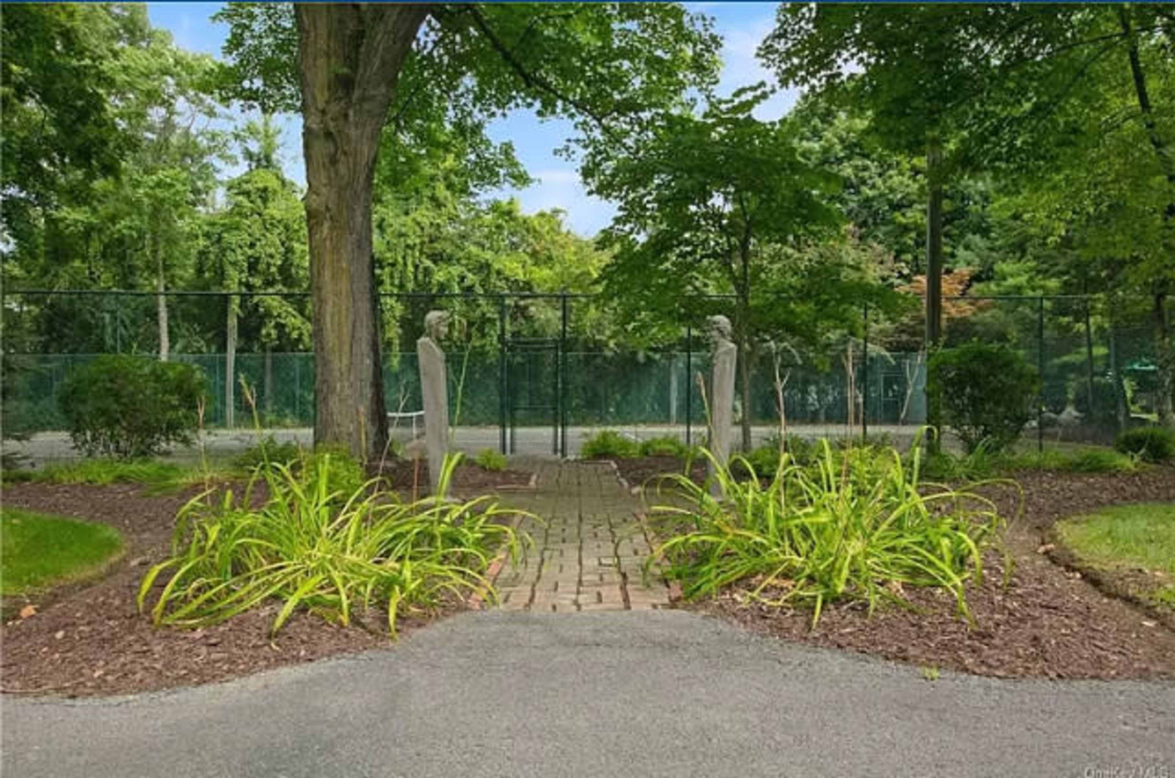 A pathway lined with plants leading to a fenced tennis court in a park-like setting.