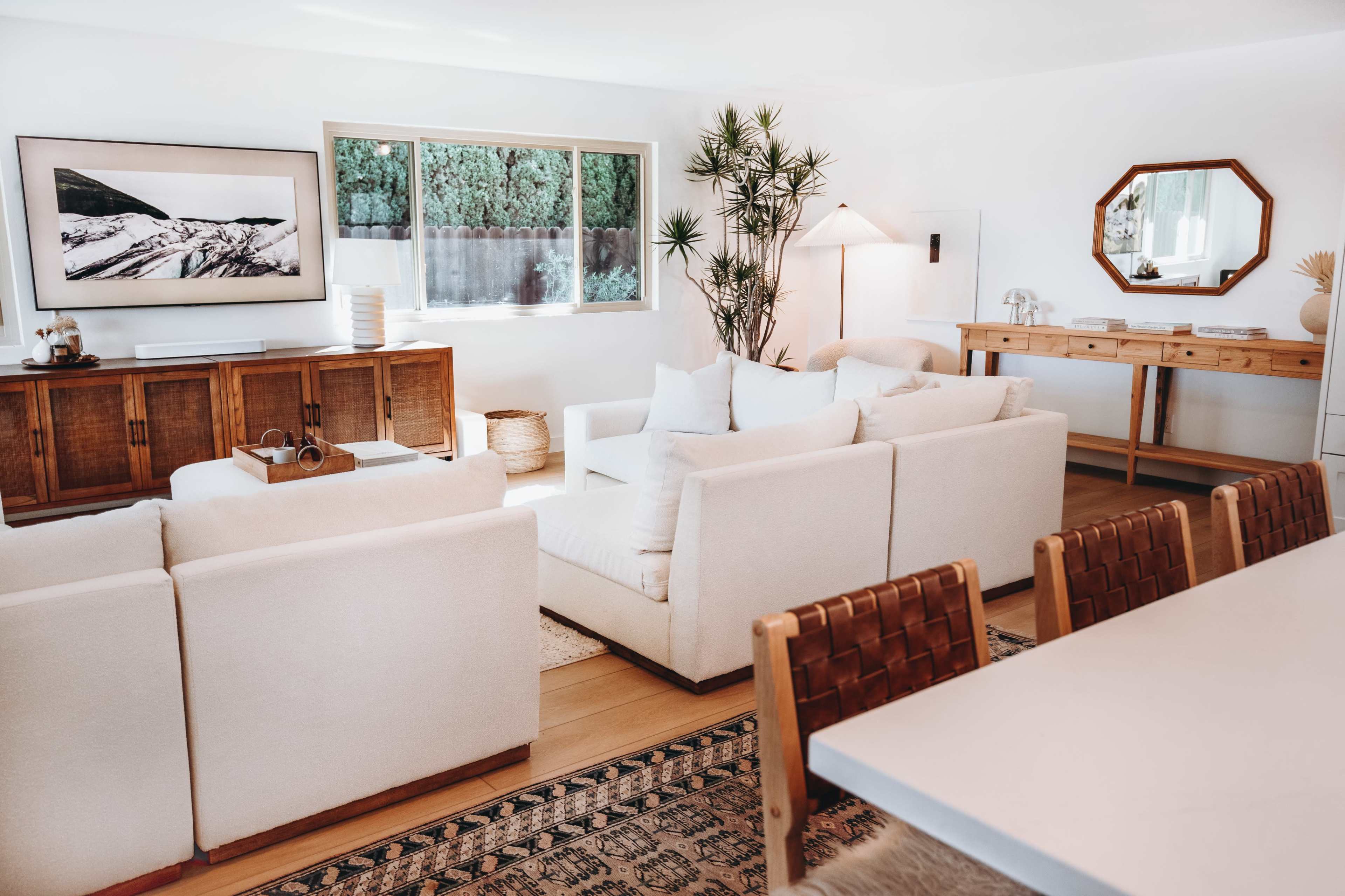 A bright, modern living room with white furniture, wood accents, and a large window overlooking greenery.