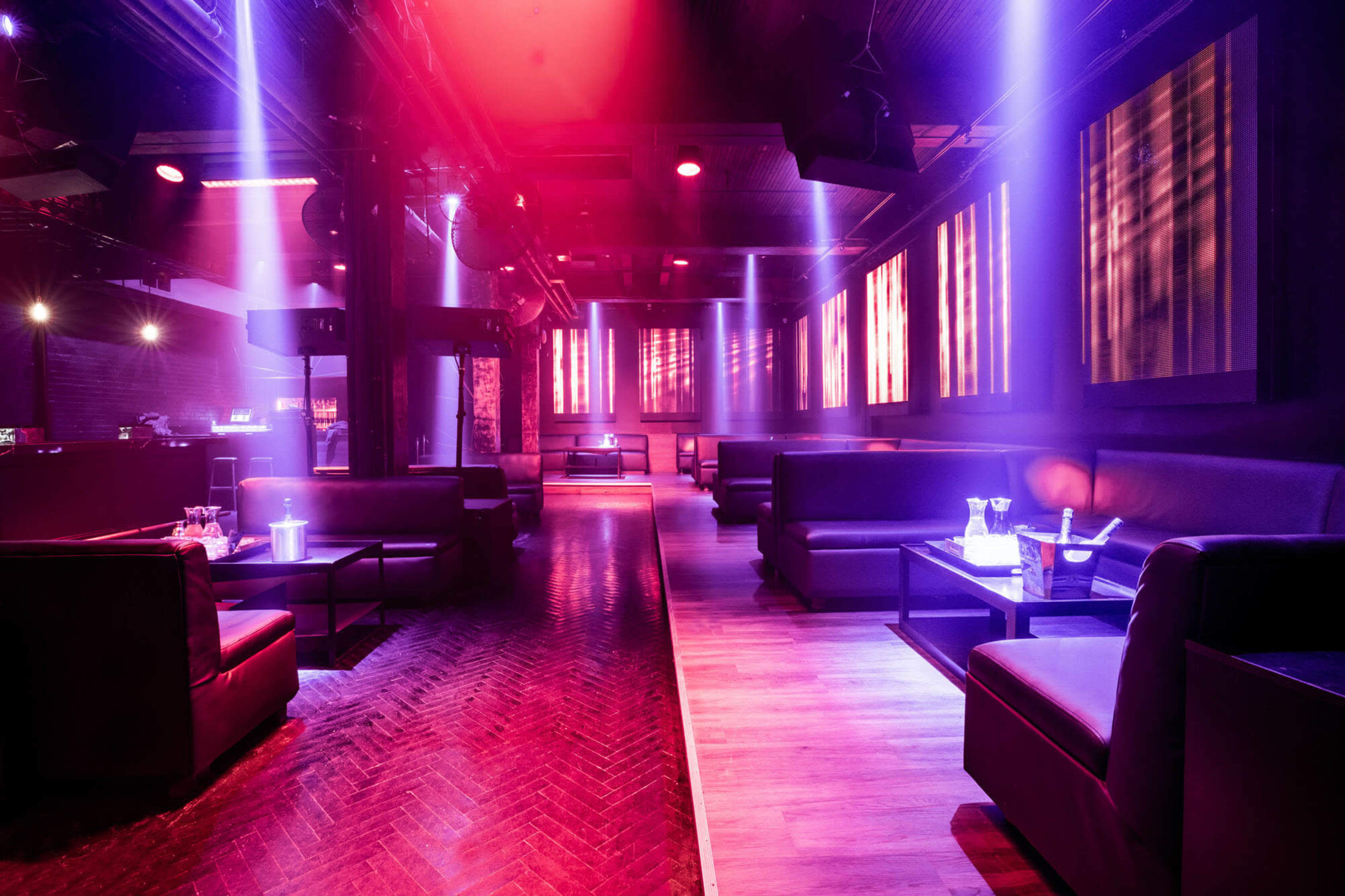 A dimly lit nightclub features sleek seating and illuminated bar areas, with colorful lights dancing across the walls.