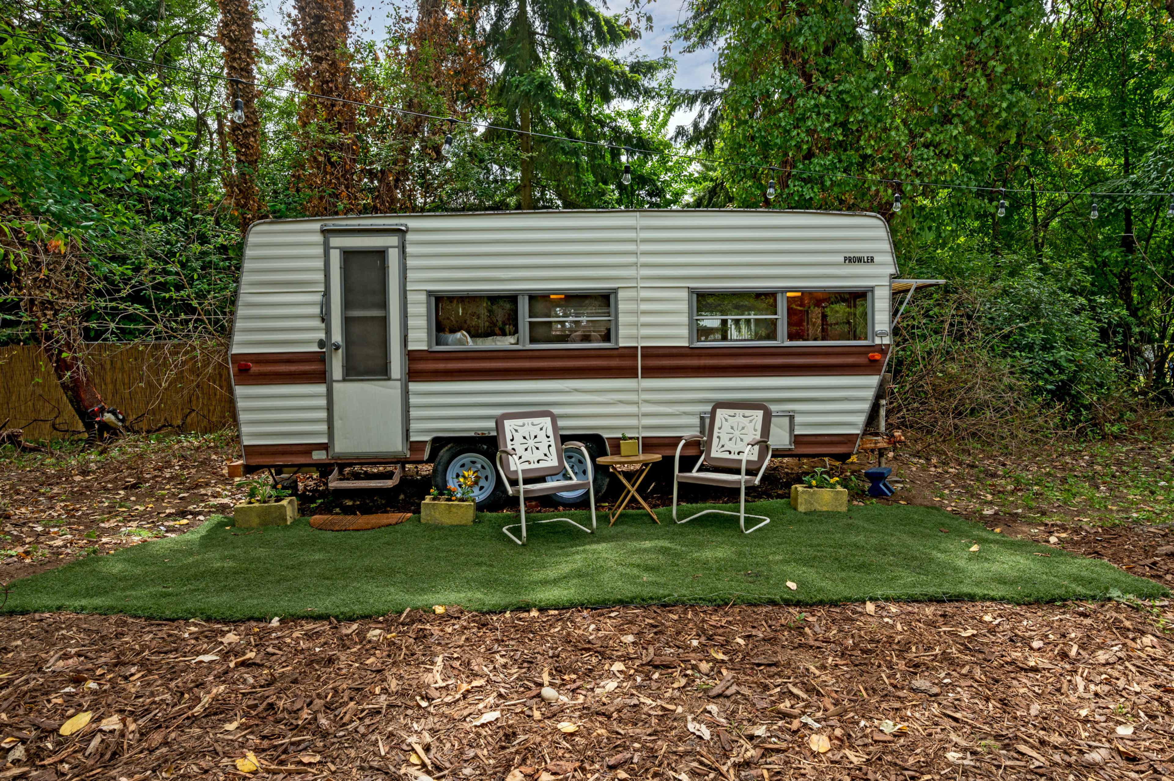 A vintage camper is parked on a patch of grass surrounded by trees, with two white chairs and a small table set in front.