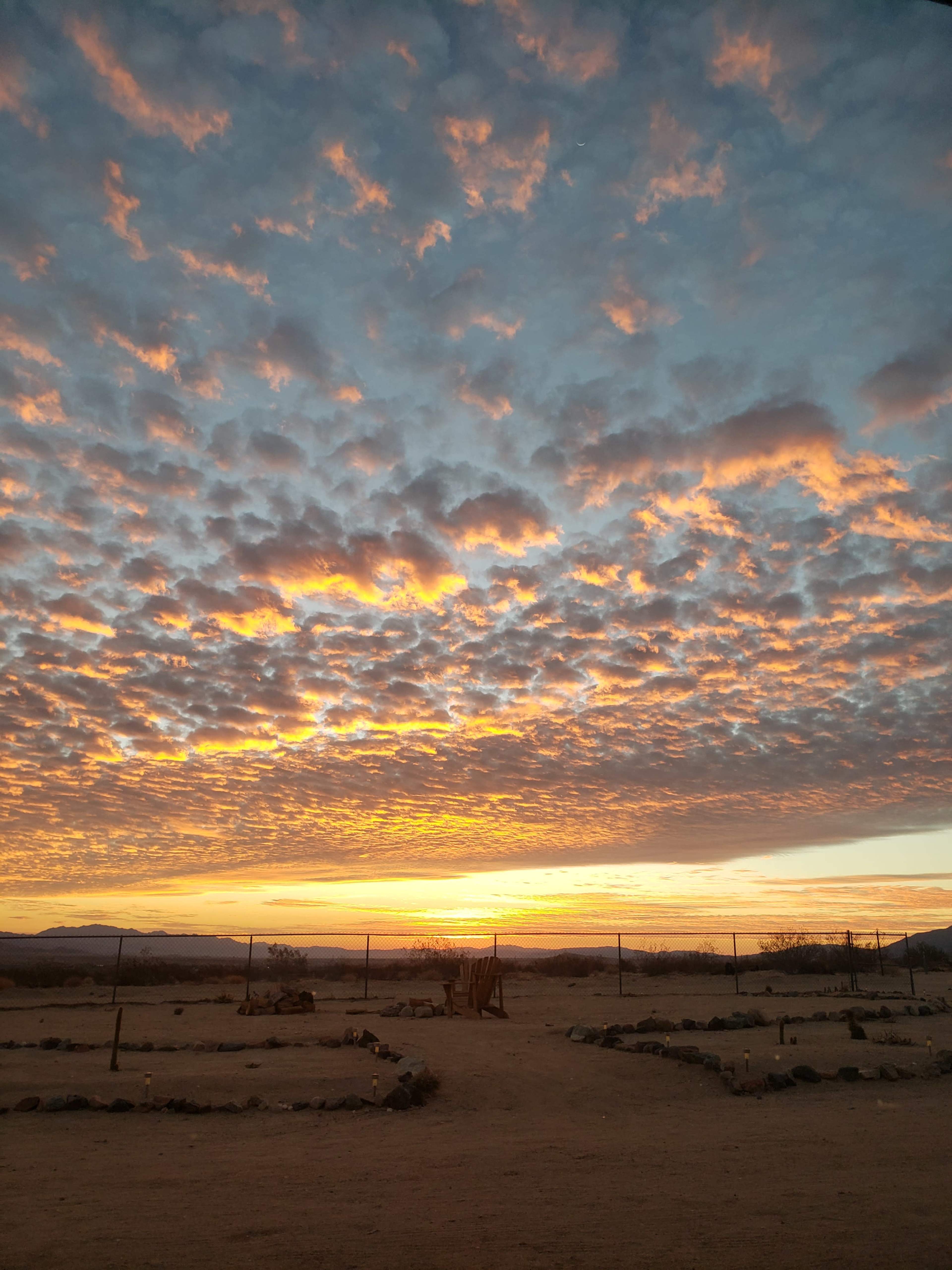 A vibrant sunset casts orange and yellow hues across a sky filled with clouds over a remote desert landscape.