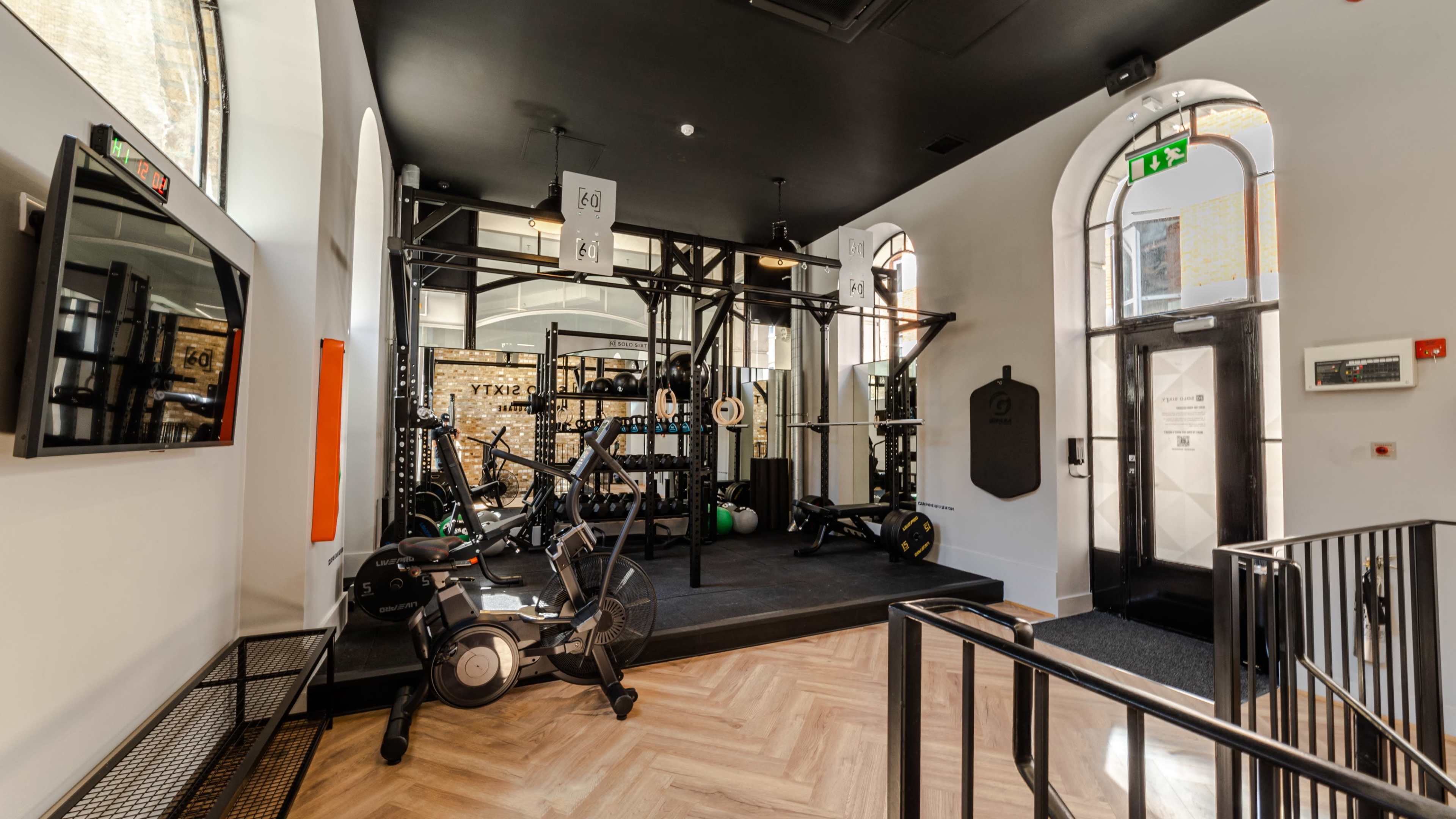 The image shows a well-equipped gym area featuring various fitness machines and weights, located in a modern, well-lit space with architectural details.