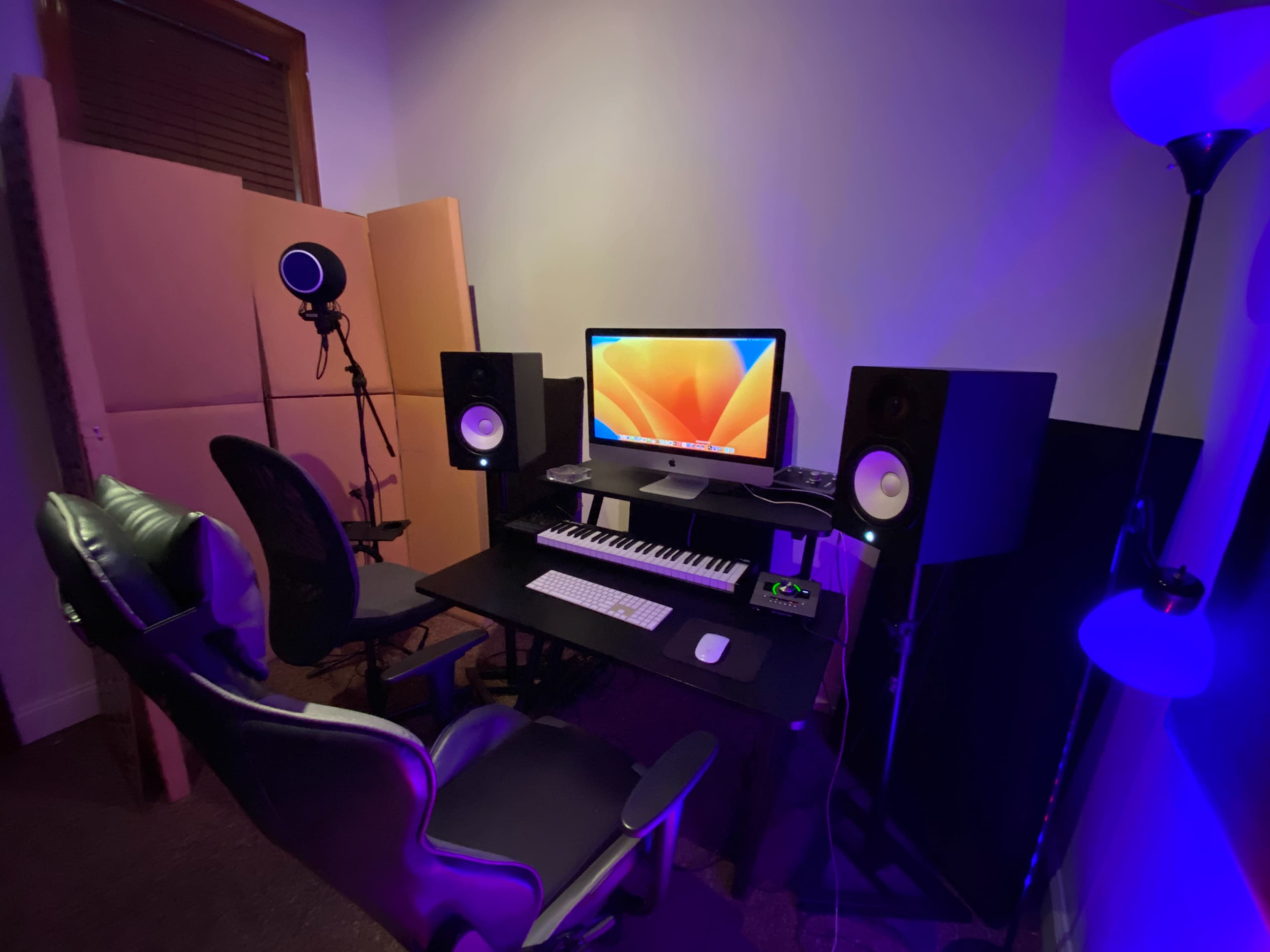 The image shows a music production setup featuring a computer, keyboard, two speakers, a microphone, and a chair, all situated in a dimly lit room.