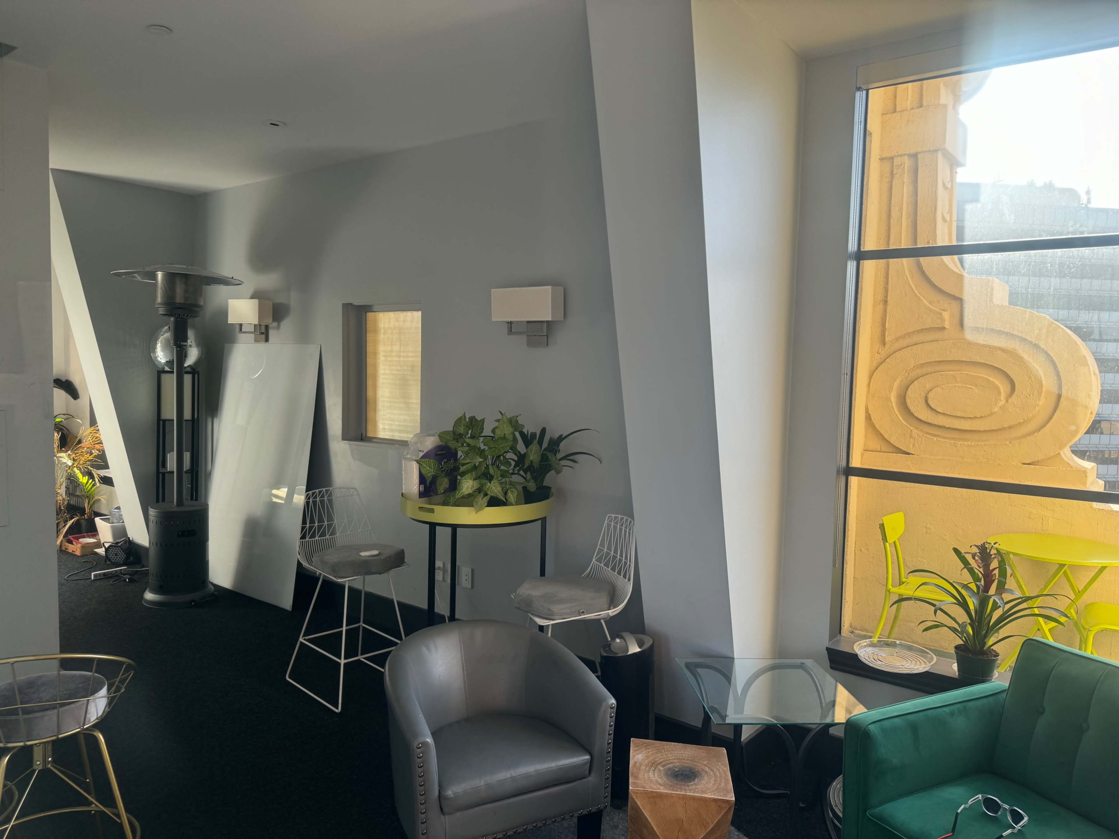 A modern interior space featuring a seating area with plants, a bar-like table, and large windows with decorative architectural details.
