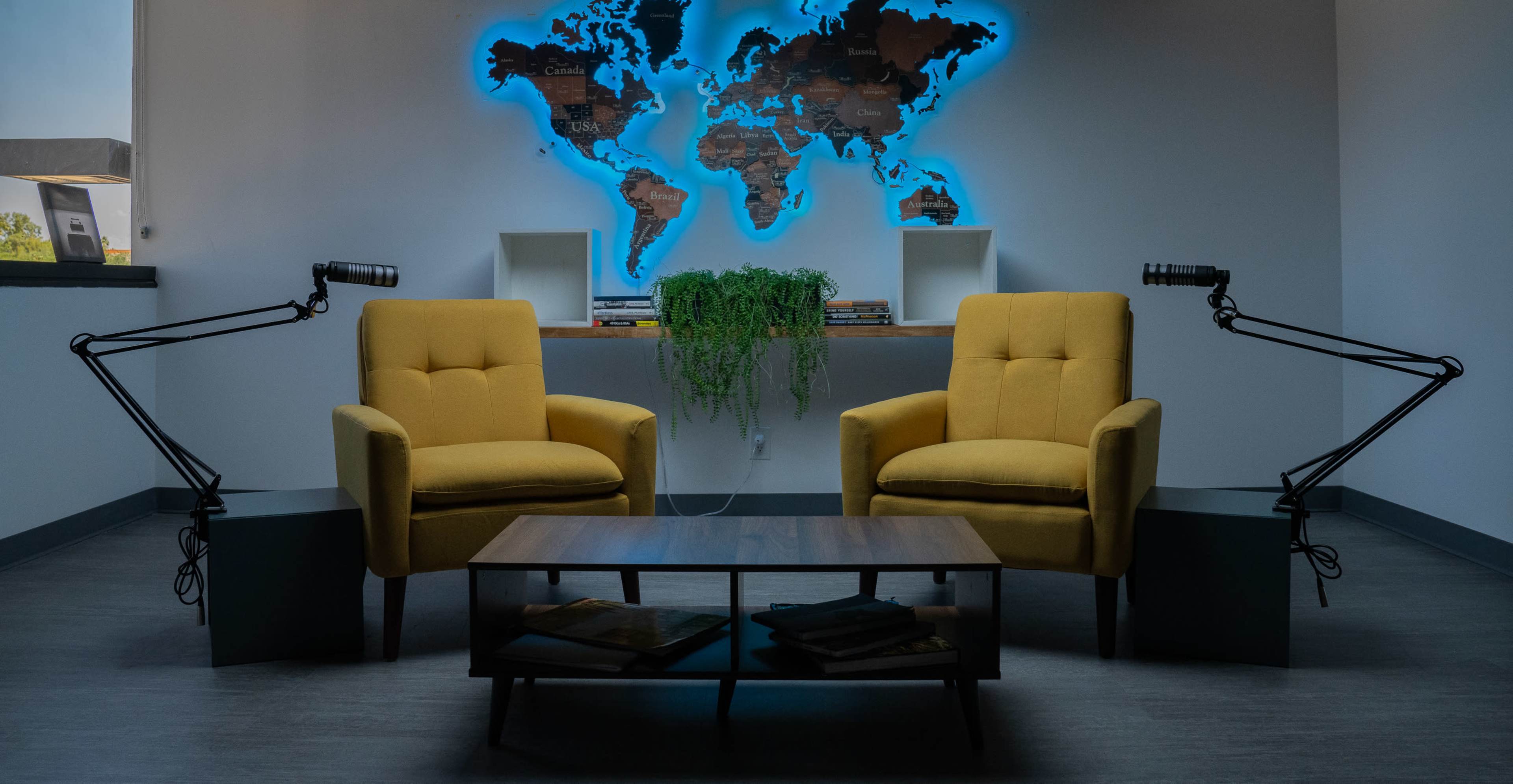 The image shows two yellow chairs facing each other in a room with a world map illuminated in blue on the wall behind them, accompanied by a coffee table and a hanging plant.