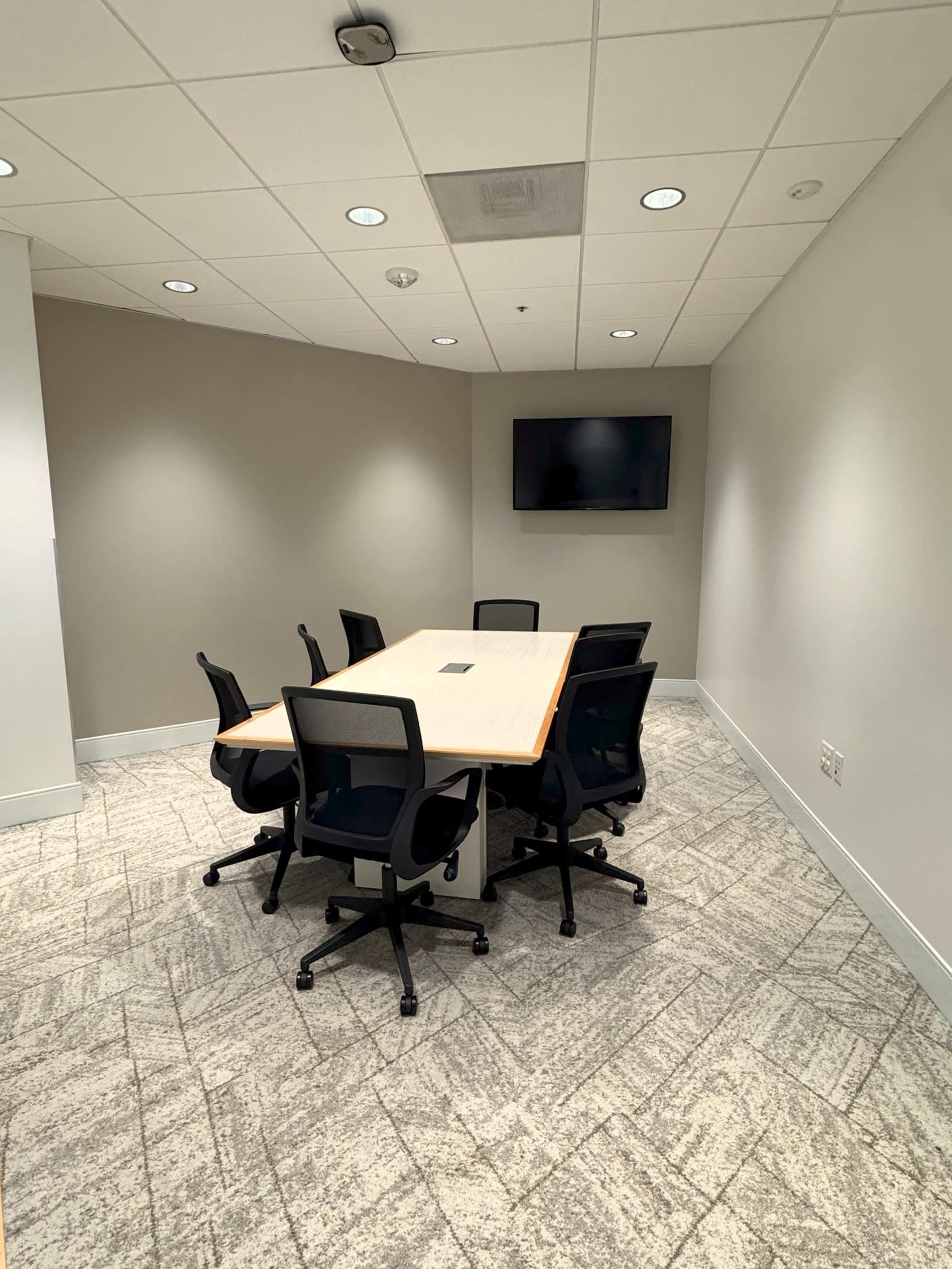 The image shows a small conference room with a rectangular wooden table surrounded by six black chairs and a wall-mounted television.