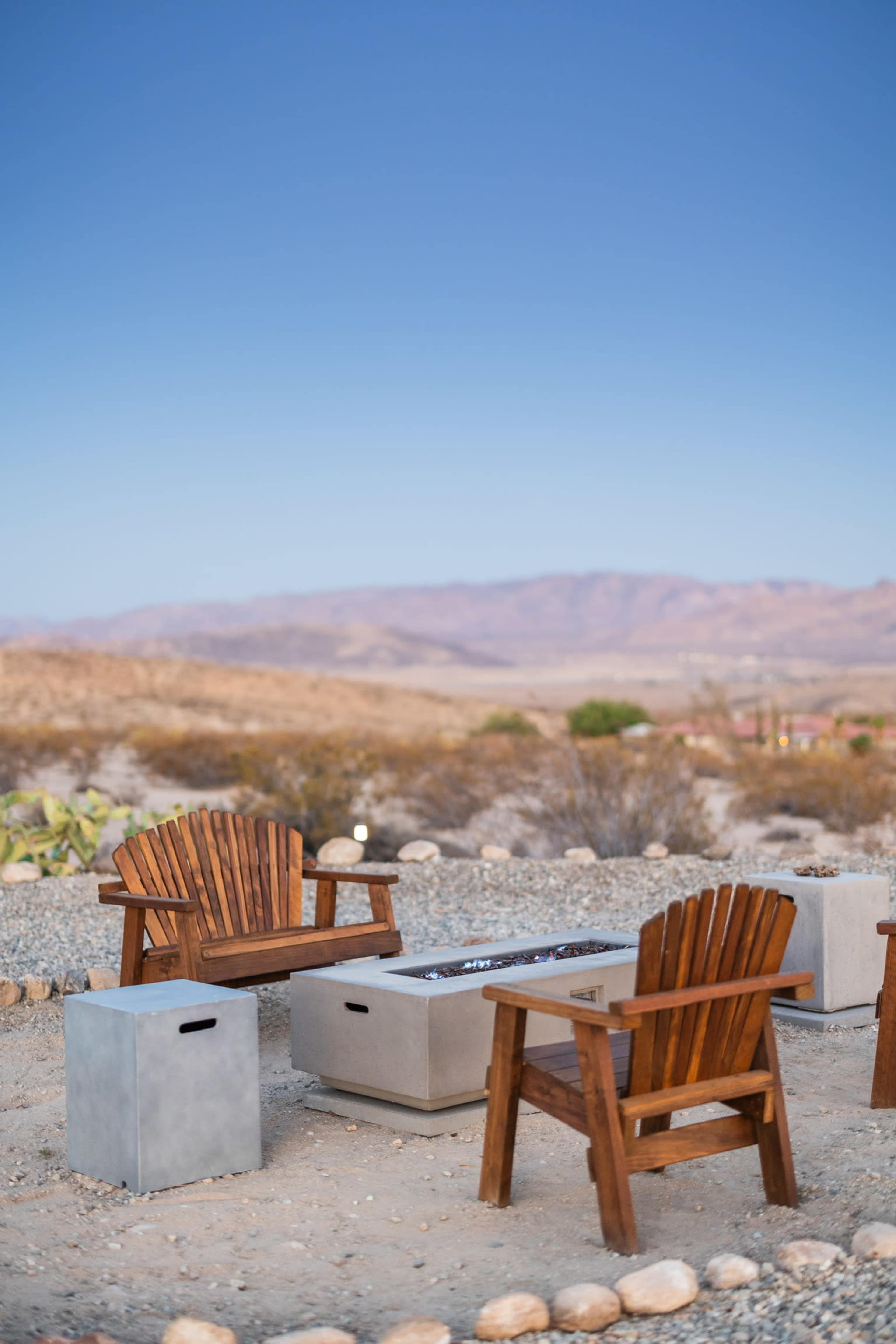 Two wooden Adirondack chairs are positioned around a rectangular fire pit on a gravel area with mountains in the background.