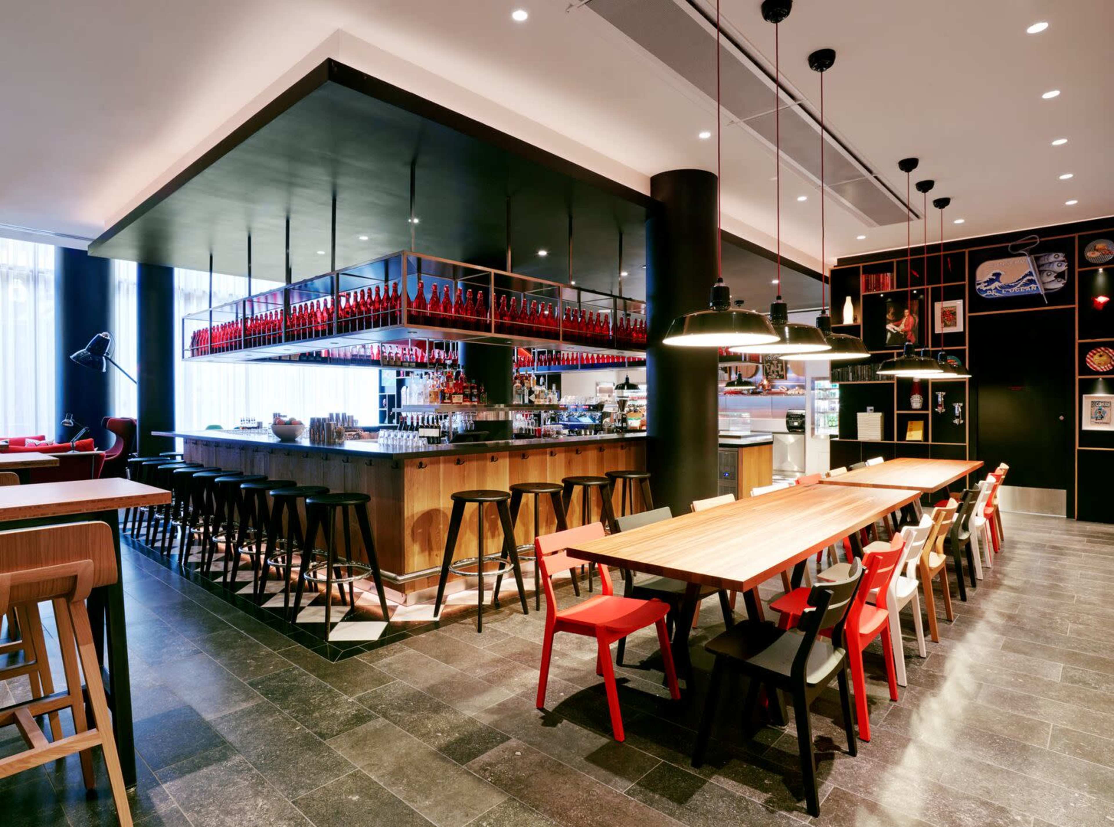 A modern restaurant interior featuring a long communal table with red and black chairs, a bar area with high stools, and shelves displaying various bottles and decorations.
