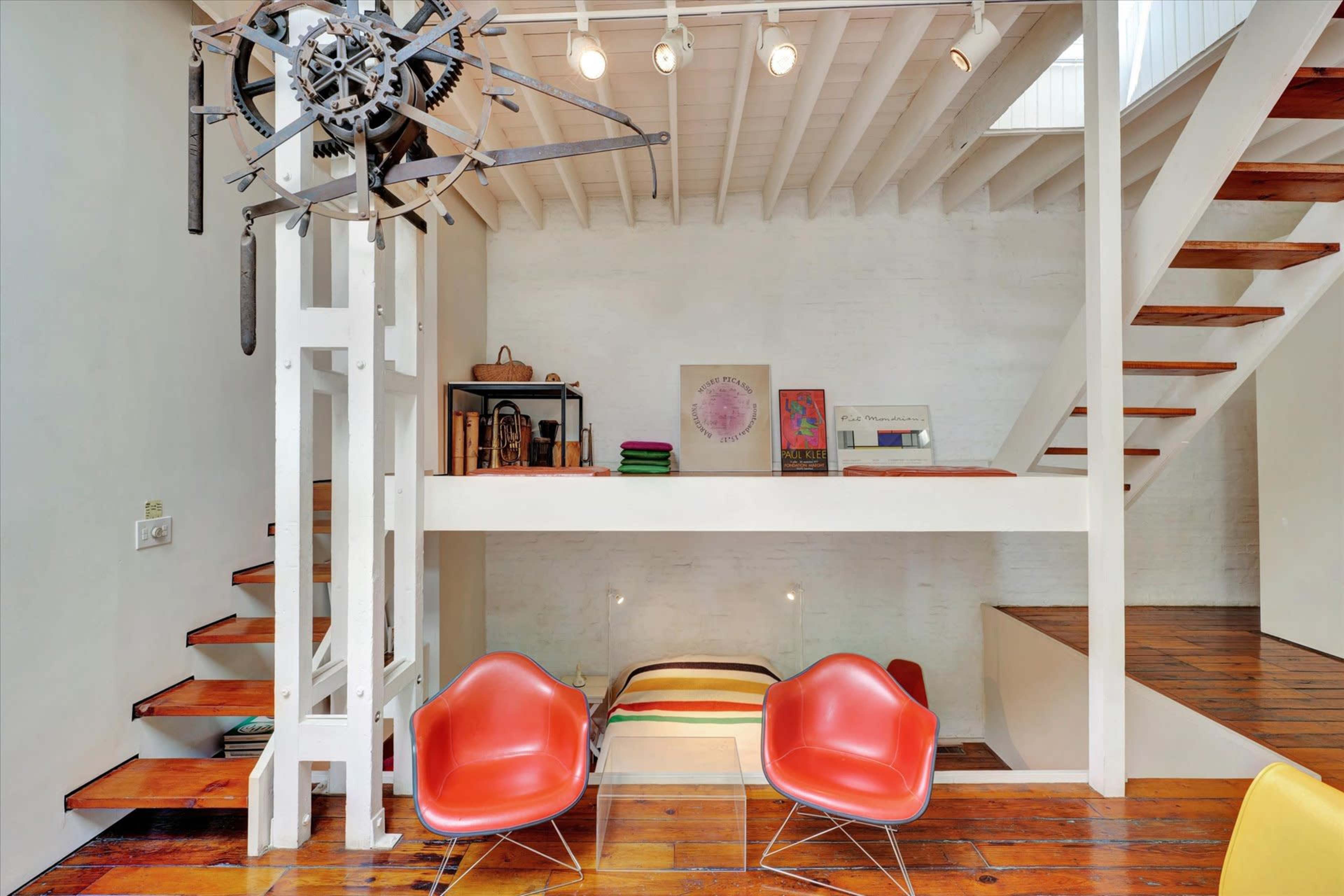A modern interior features a wooden staircase, a lofted area with a bed, and two red chairs under a mechanical clock installation.