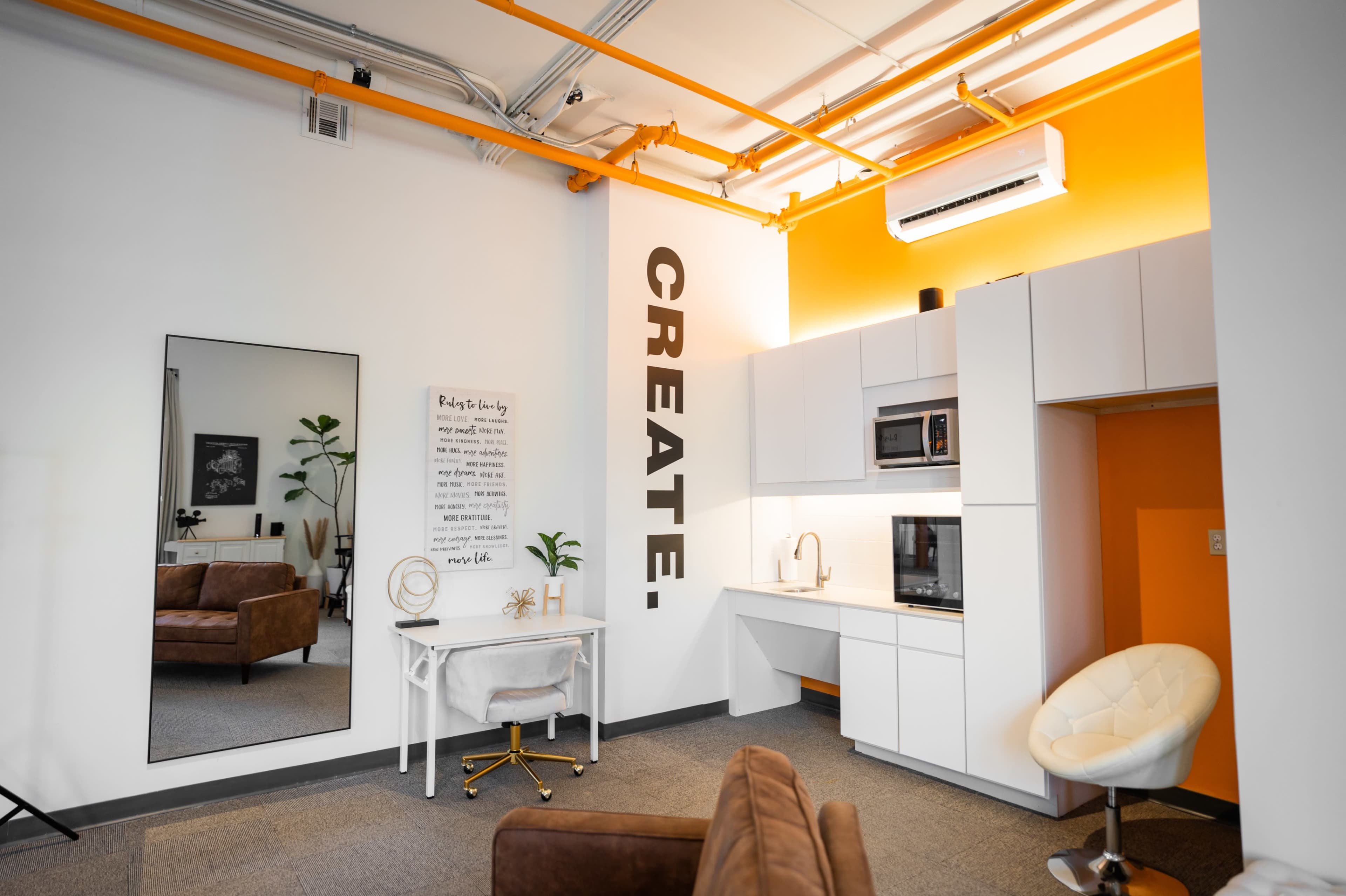 A modern workspace with a kitchen area, a mirror, and a wall featuring the word "CREATE." in large letters.