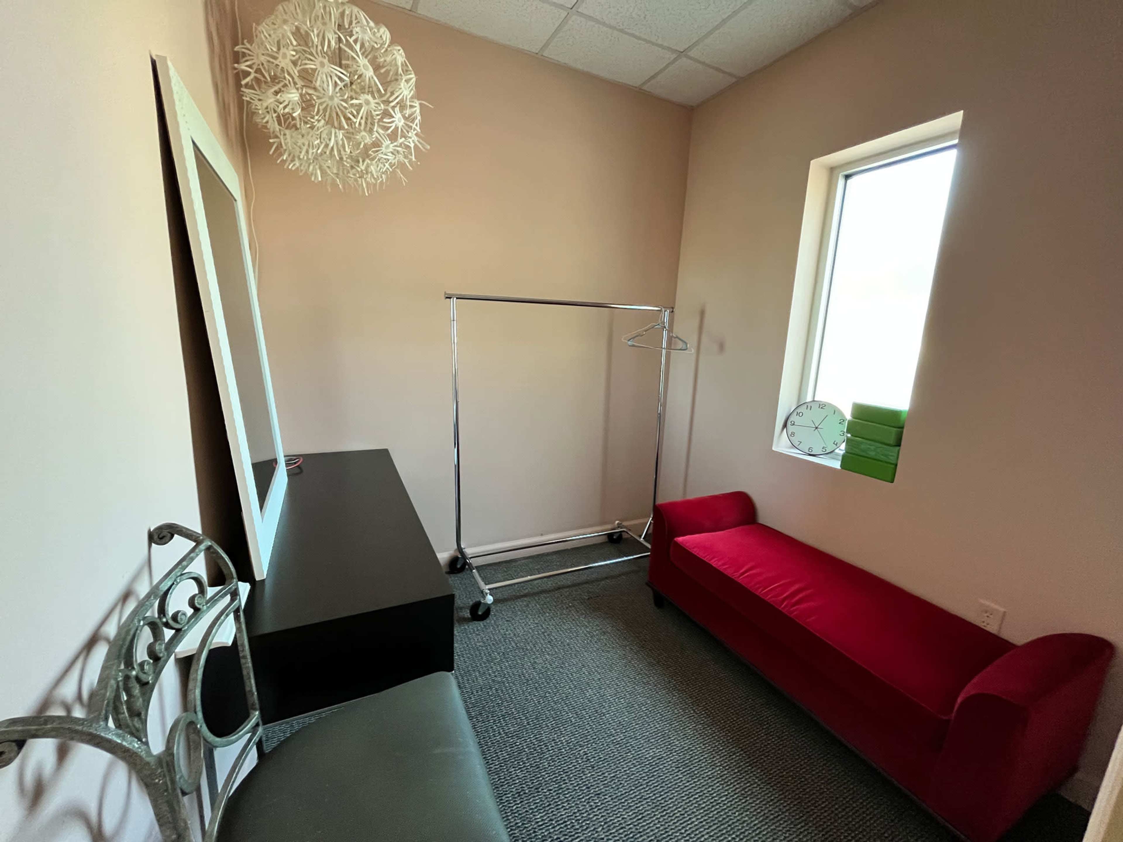 A small room with a pink wall, a red couch, a clothing rack, a large mirror, and a clock on a table.