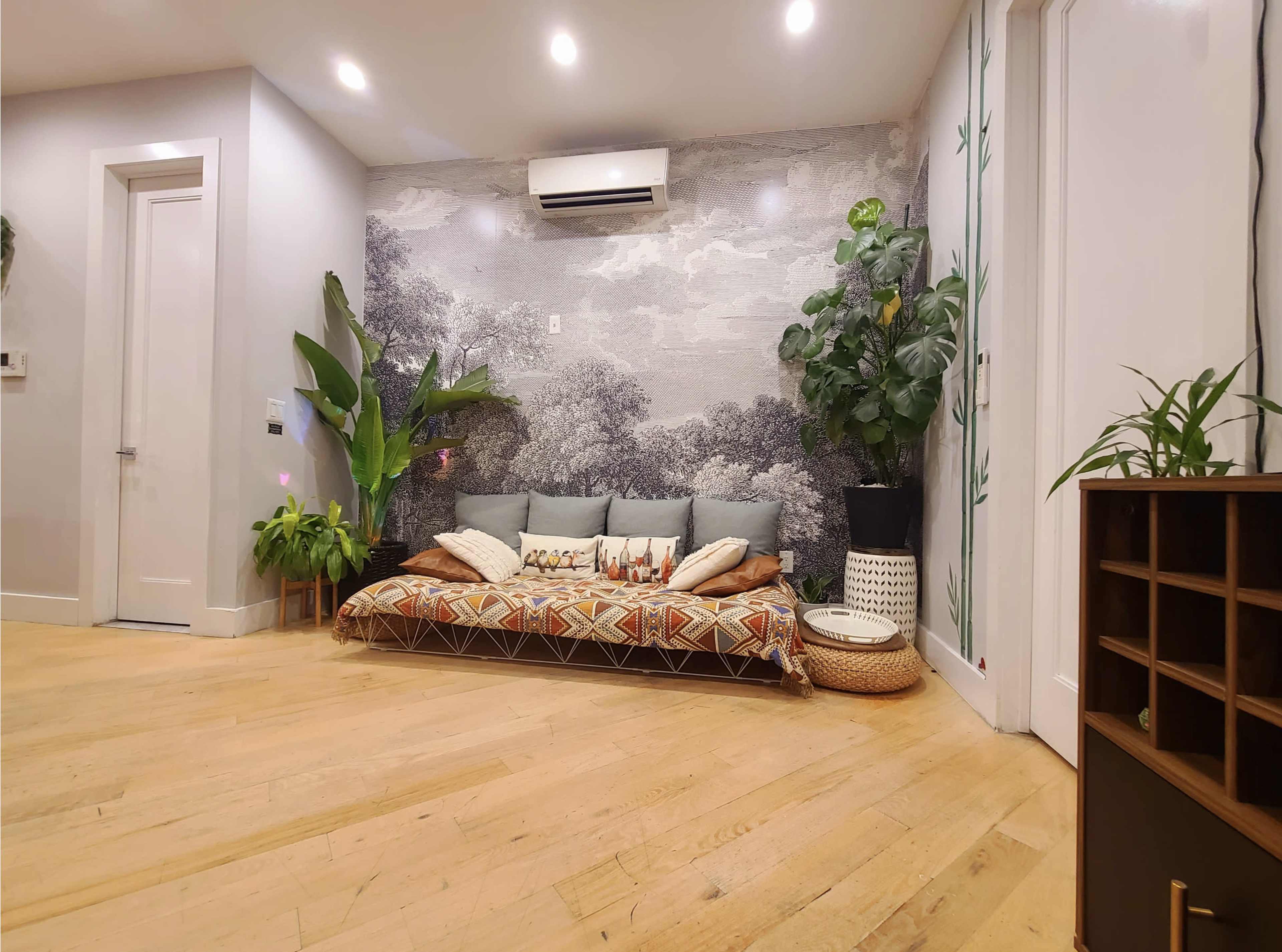 The room features a low seating area with cushions against a mural of trees, surrounded by potted plants and wooden flooring.