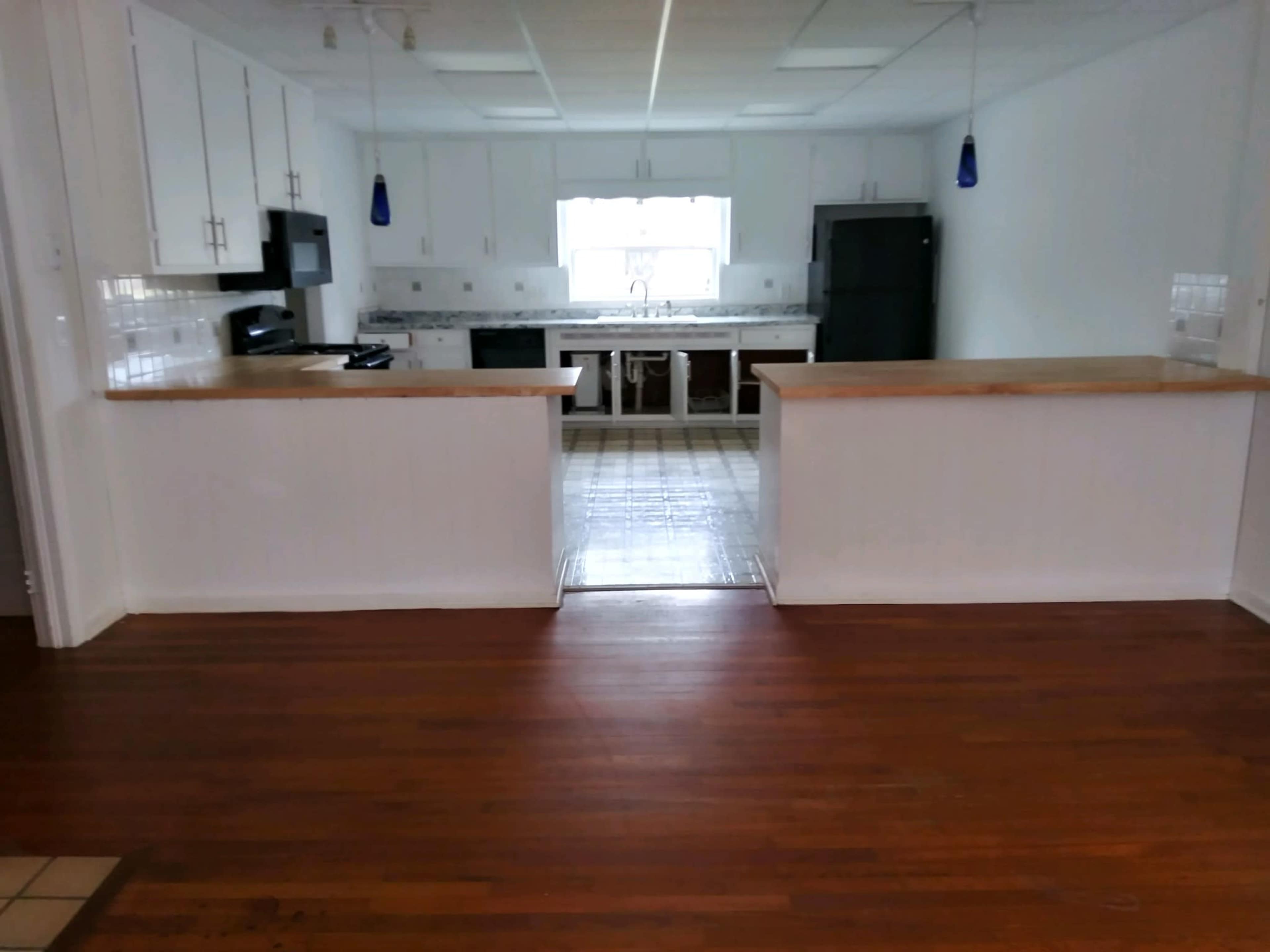 The image shows a kitchen with white cabinets, a central island, and wooden flooring, featuring a window providing natural light.