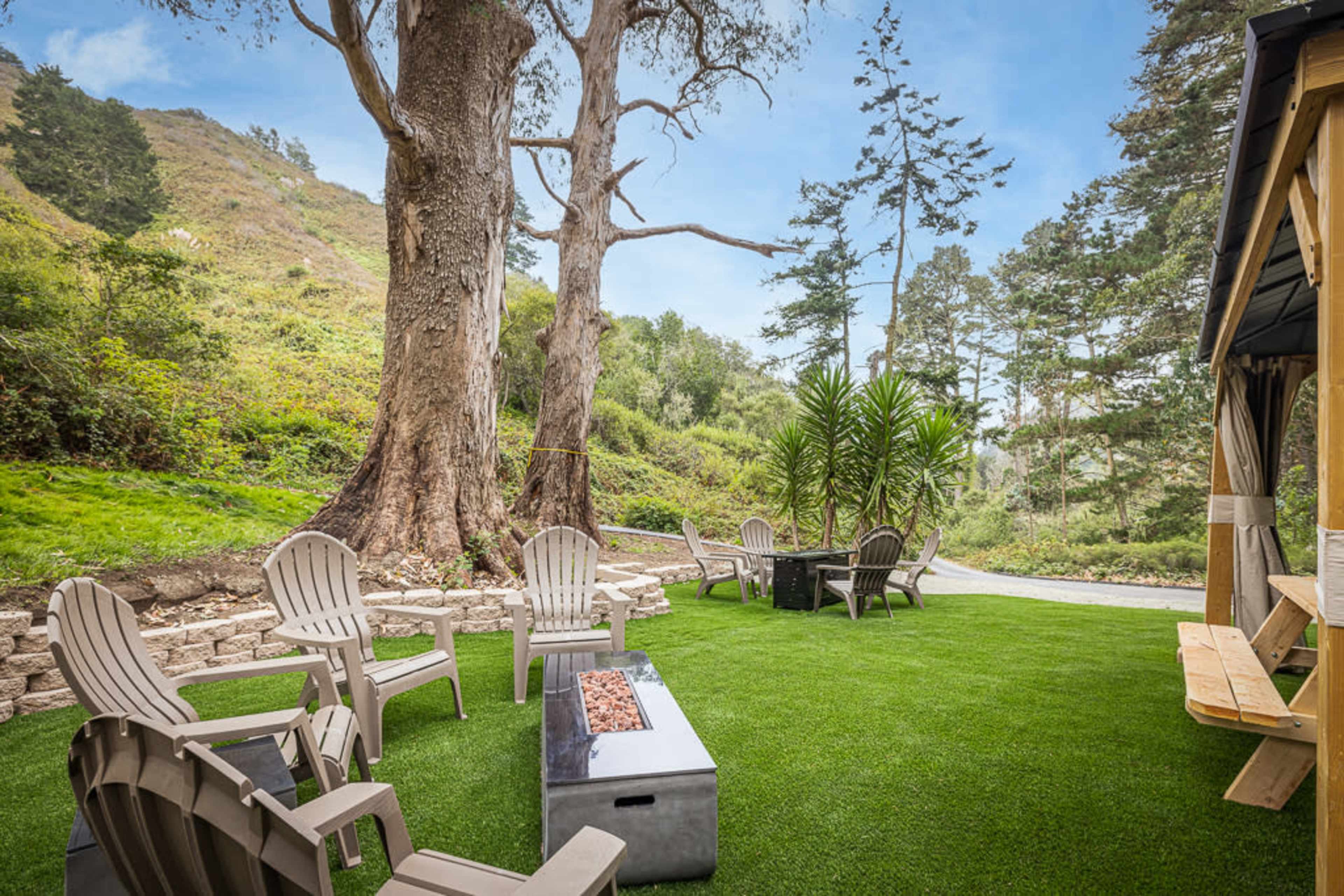 A patio area with several Adirondack chairs surrounding a fire pit, set against a backdrop of trees and a hillside.