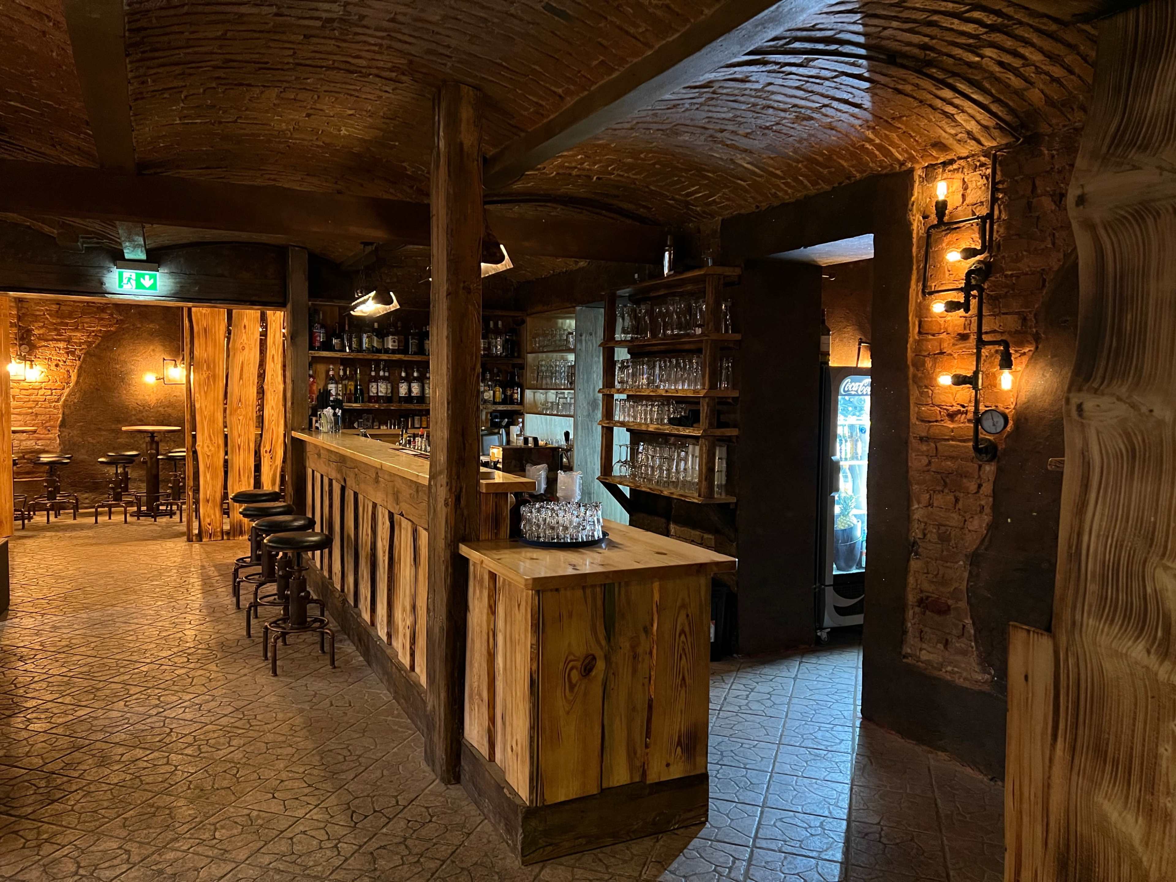 A dimly lit bar interior featuring a wooden counter, shelves lined with glassware, and seating against the walls.