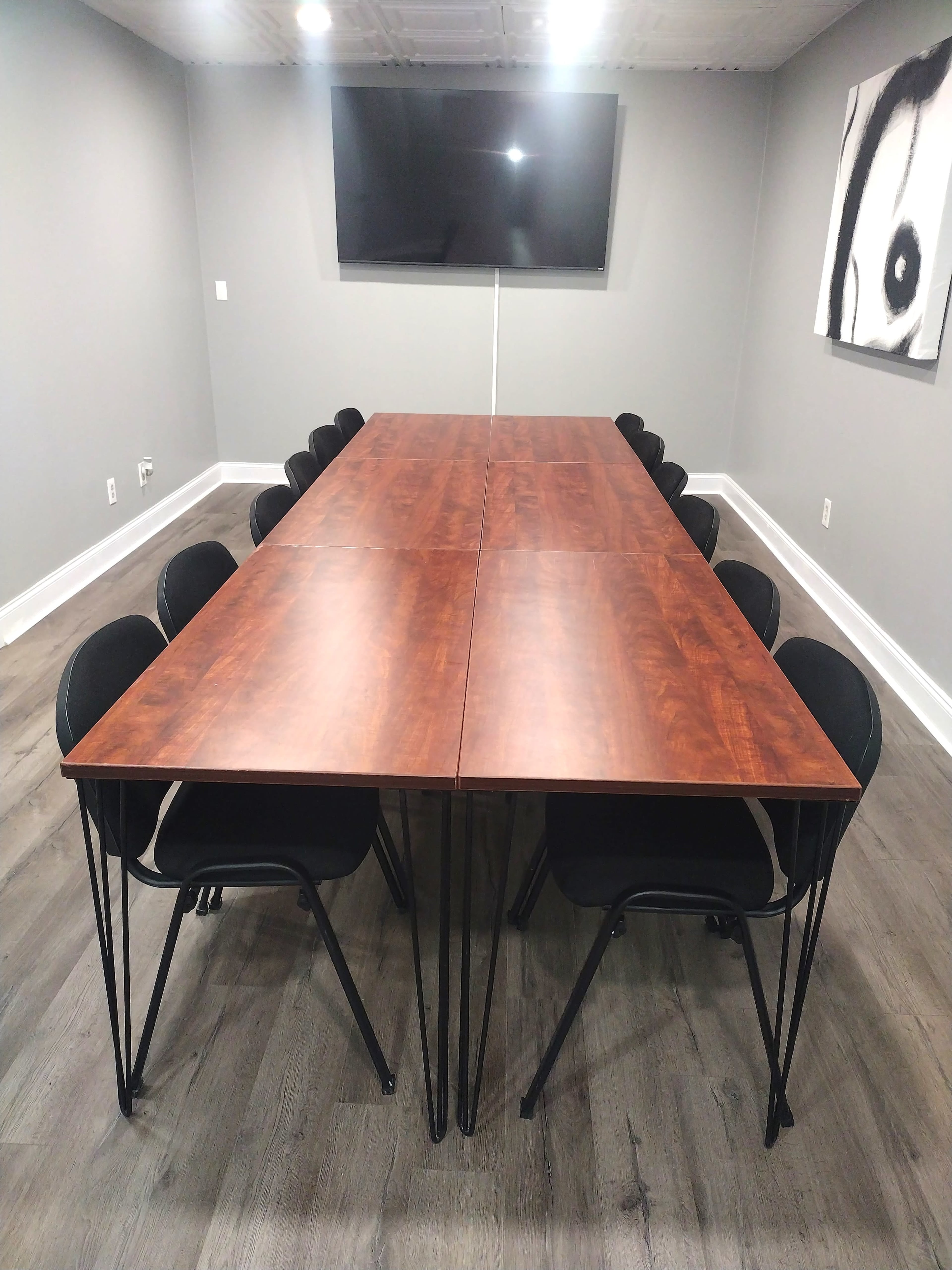 A long wooden conference table with black chairs is arranged in a meeting room featuring gray walls and a large wall-mounted television.