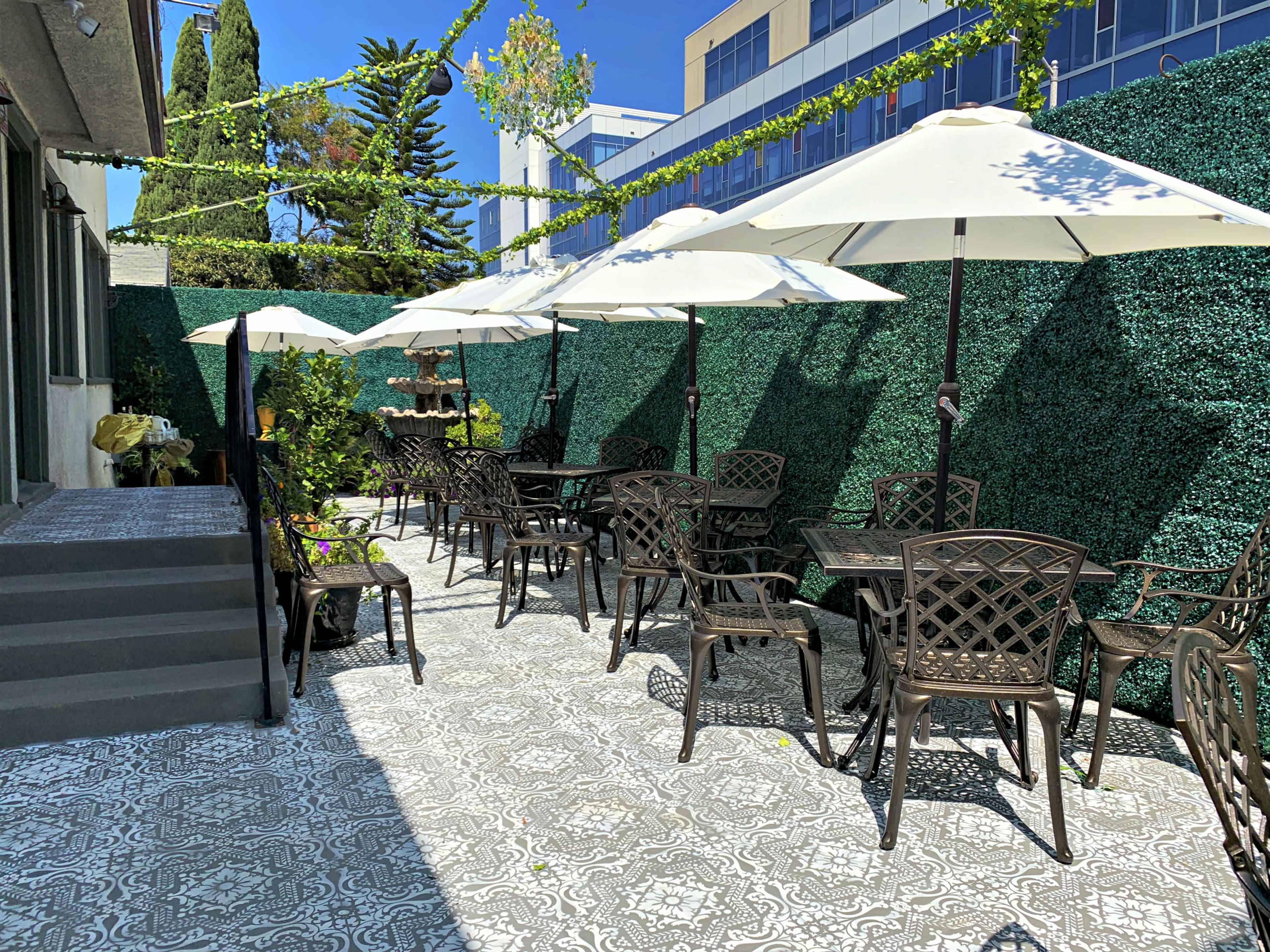 The image shows a patio area with several tables and chairs under white umbrellas, surrounded by green hedges and stepping stones.