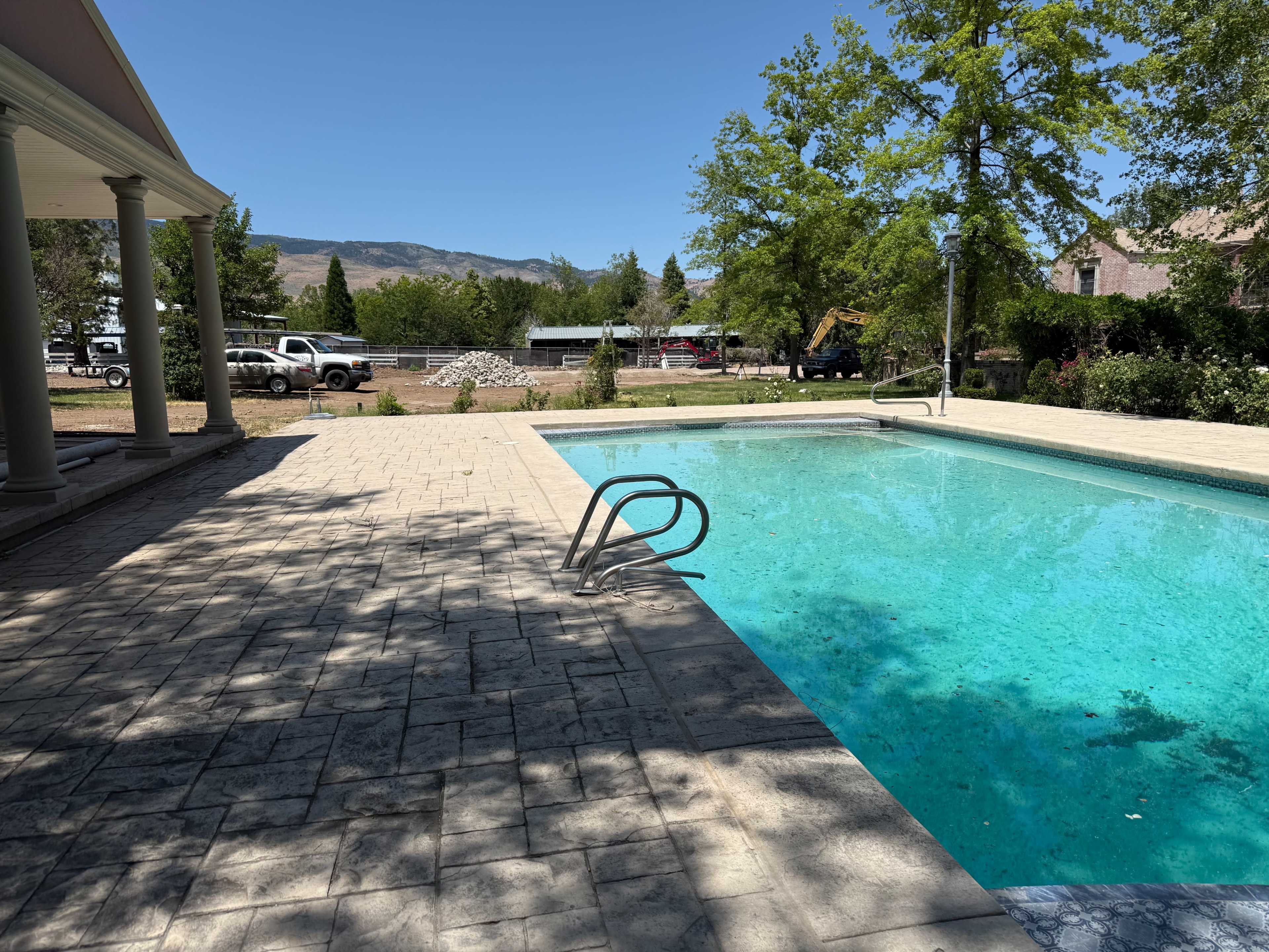 The image shows a clear swimming pool next to a patio area, with trees and vehicles in the background.
