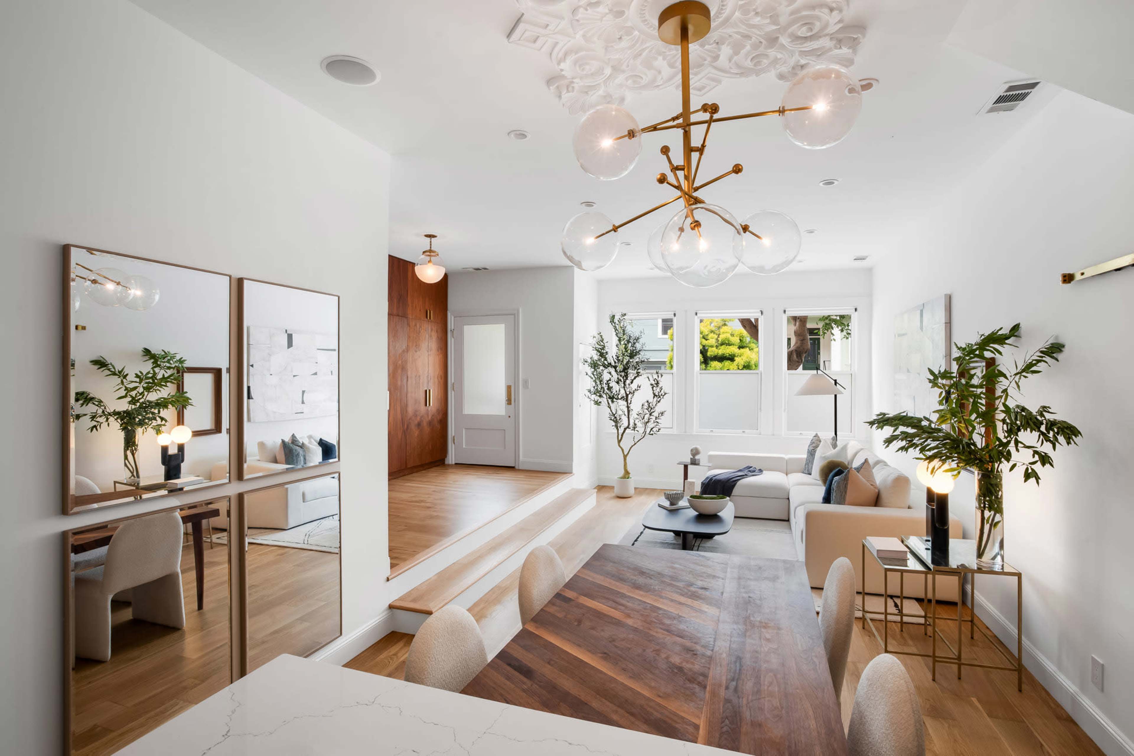 The image shows a modern living space featuring a dining area with a large wooden table, a stylish chandelier, and a cozy seating area with a sofa and plants nearby.