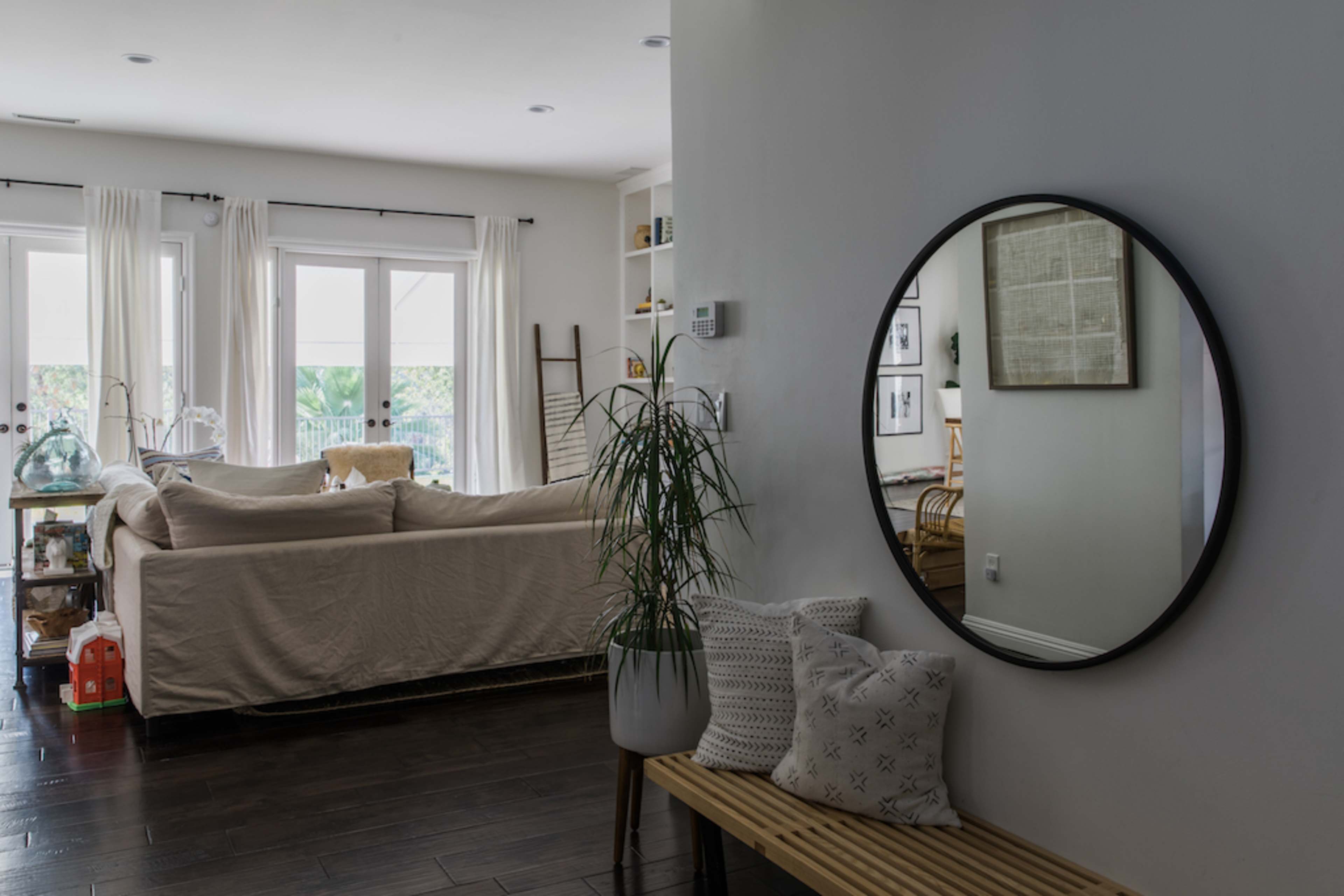 A mirror hangs on the wall opposite a living area with a beige sofa and large windows.