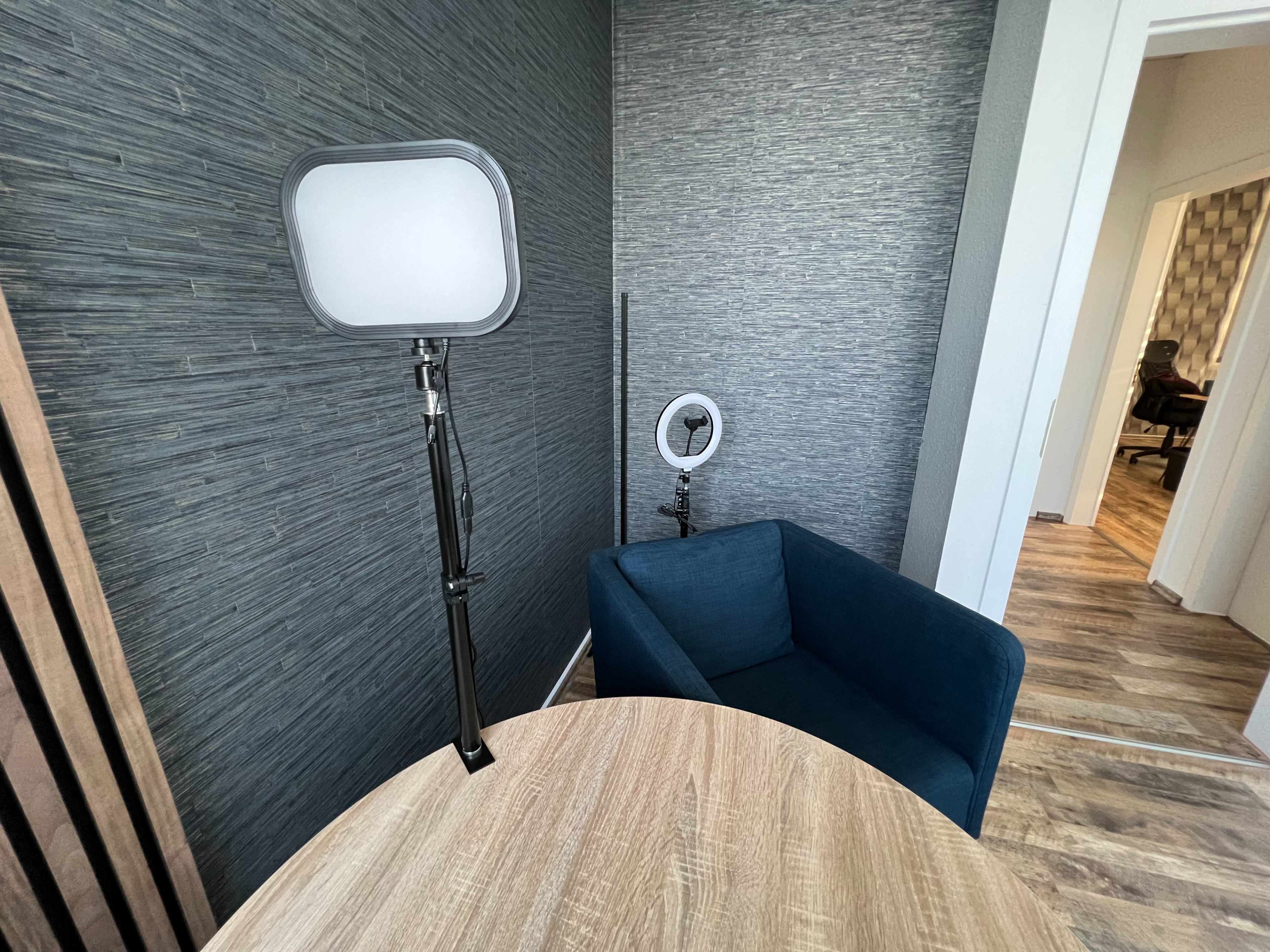 The image depicts a corner of a modern office space with a blue armchair, a wooden table, and two lamps, one of which features a ring light.