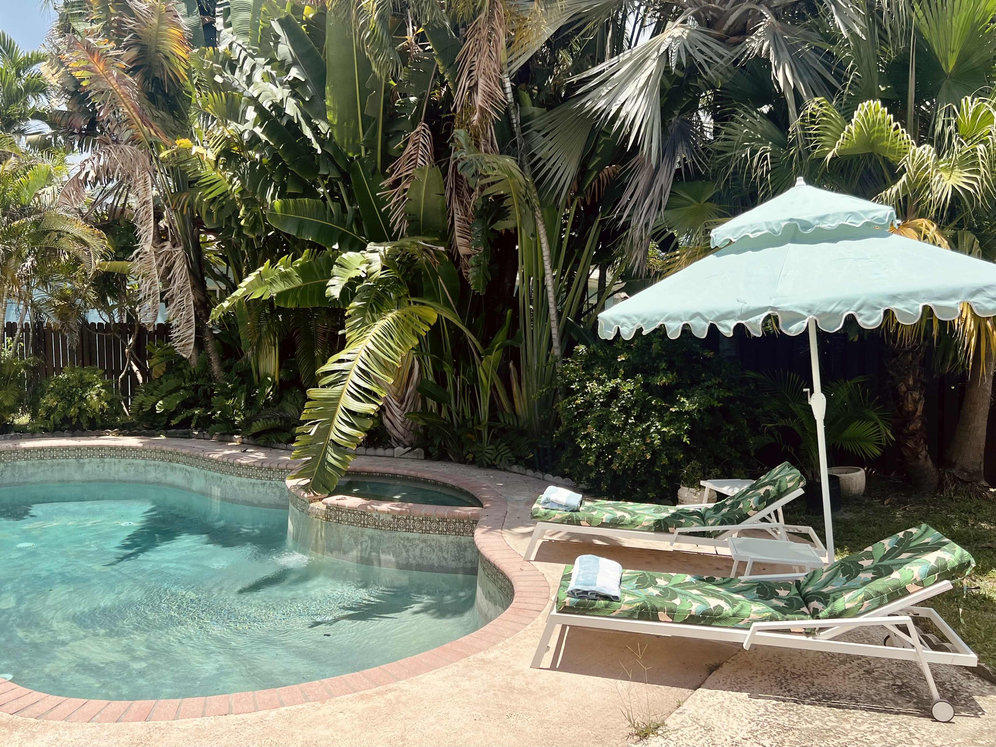 A pool area surrounded by palm trees and tropical plants, featuring two lounge chairs and a parasol.