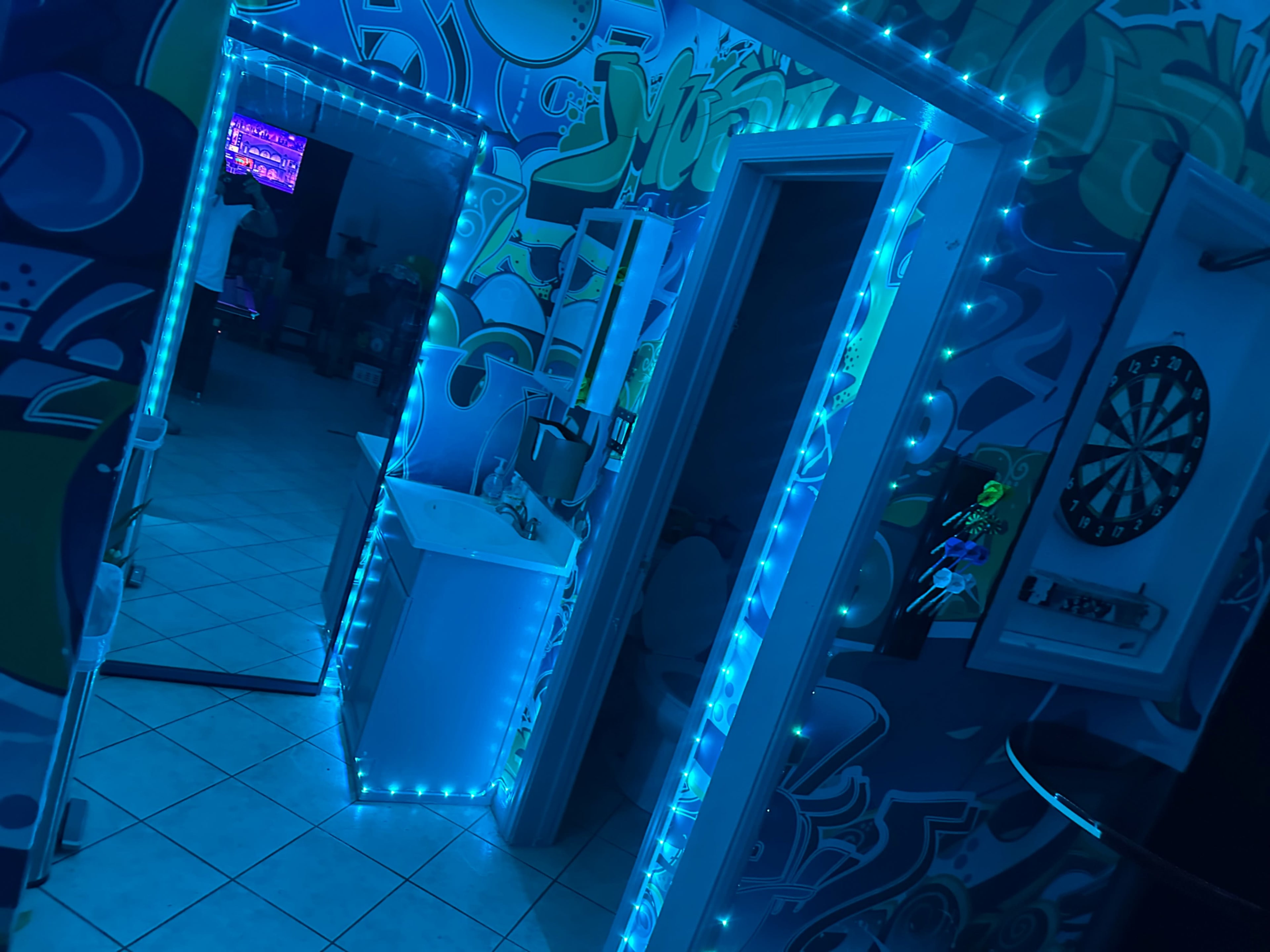 A brightly lit room features blue LED lights outlining doorways, with vibrant graffiti-style wall art and a dartboard in the background.