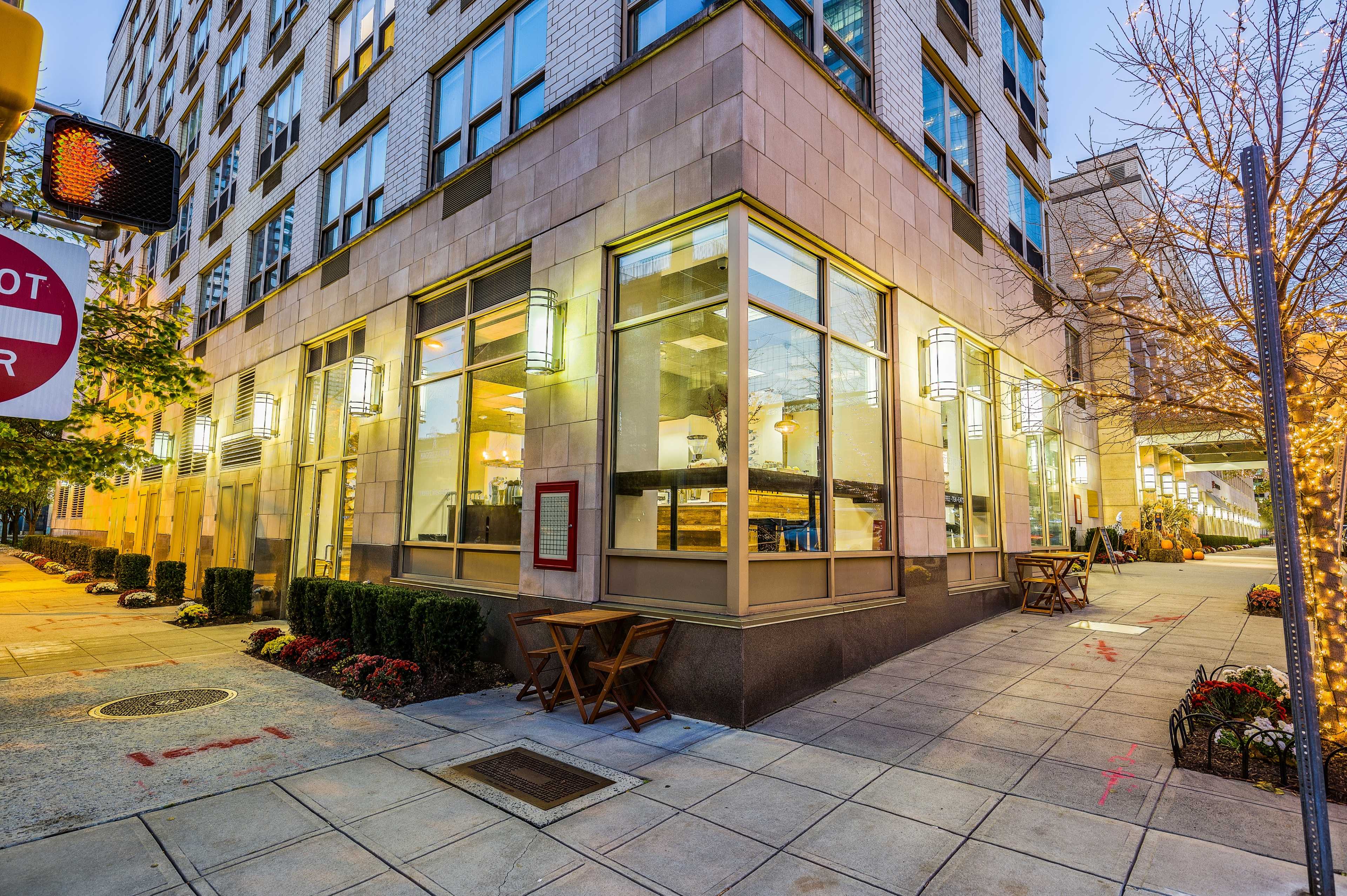 A corner view of a modern building with large windows, outdoor seating, and street lighting in the early evening.