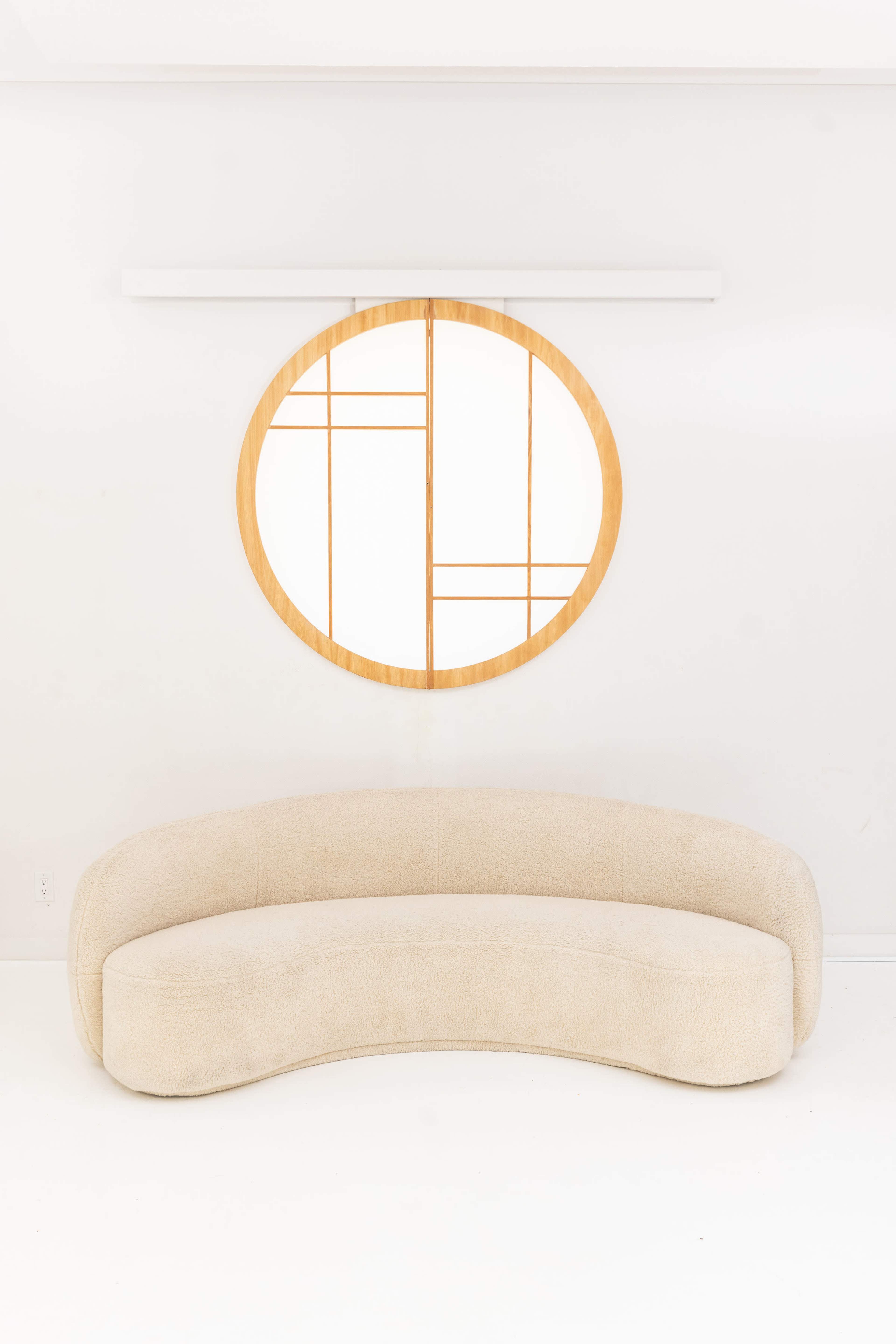 A curved, beige sofa is placed beneath a round, wooden-framed mirror with a geometric design against a white wall.