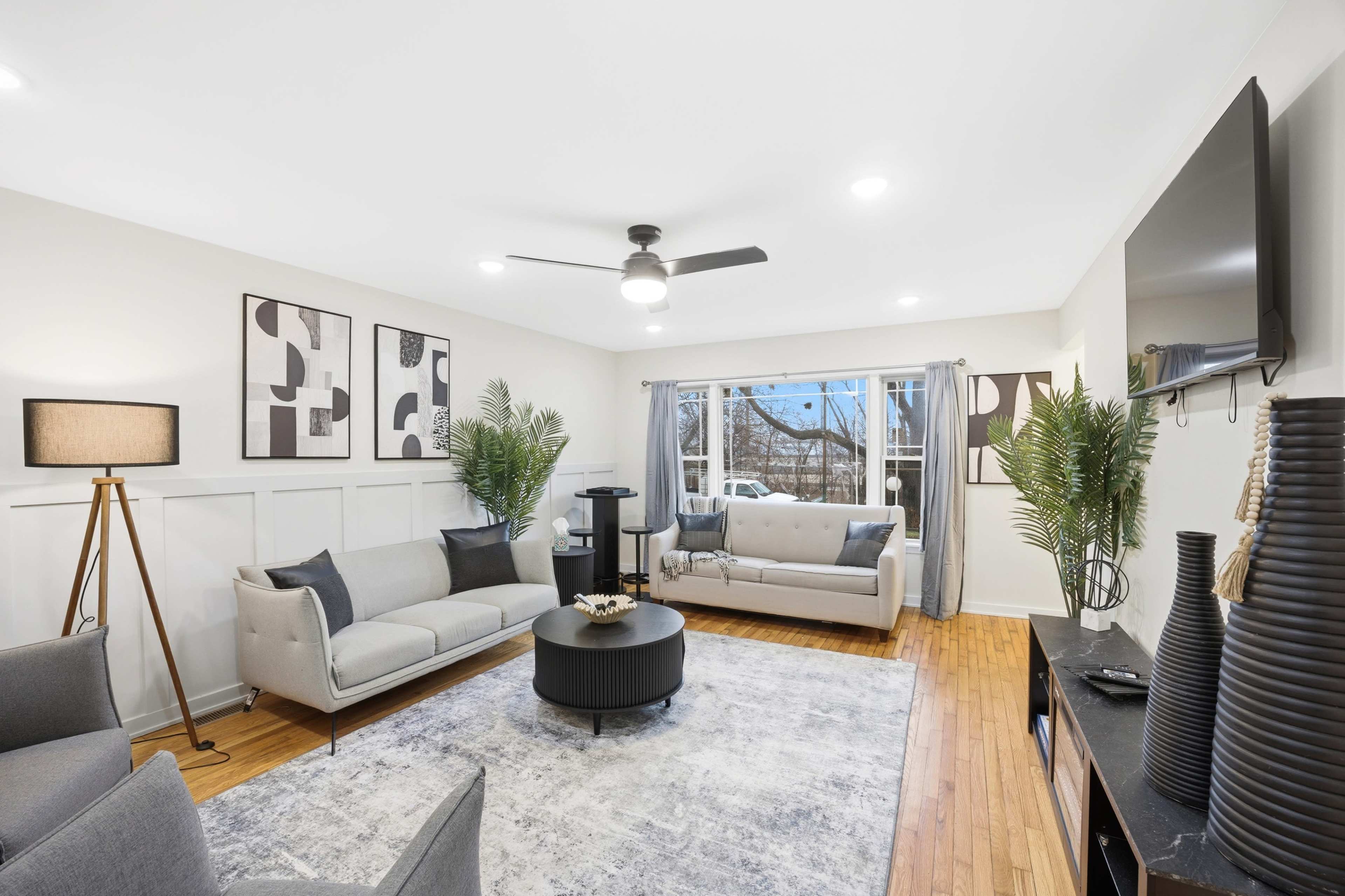 A modern living room features two sofas, a round coffee table, and large windows with curtains, surrounded by decorative plants.