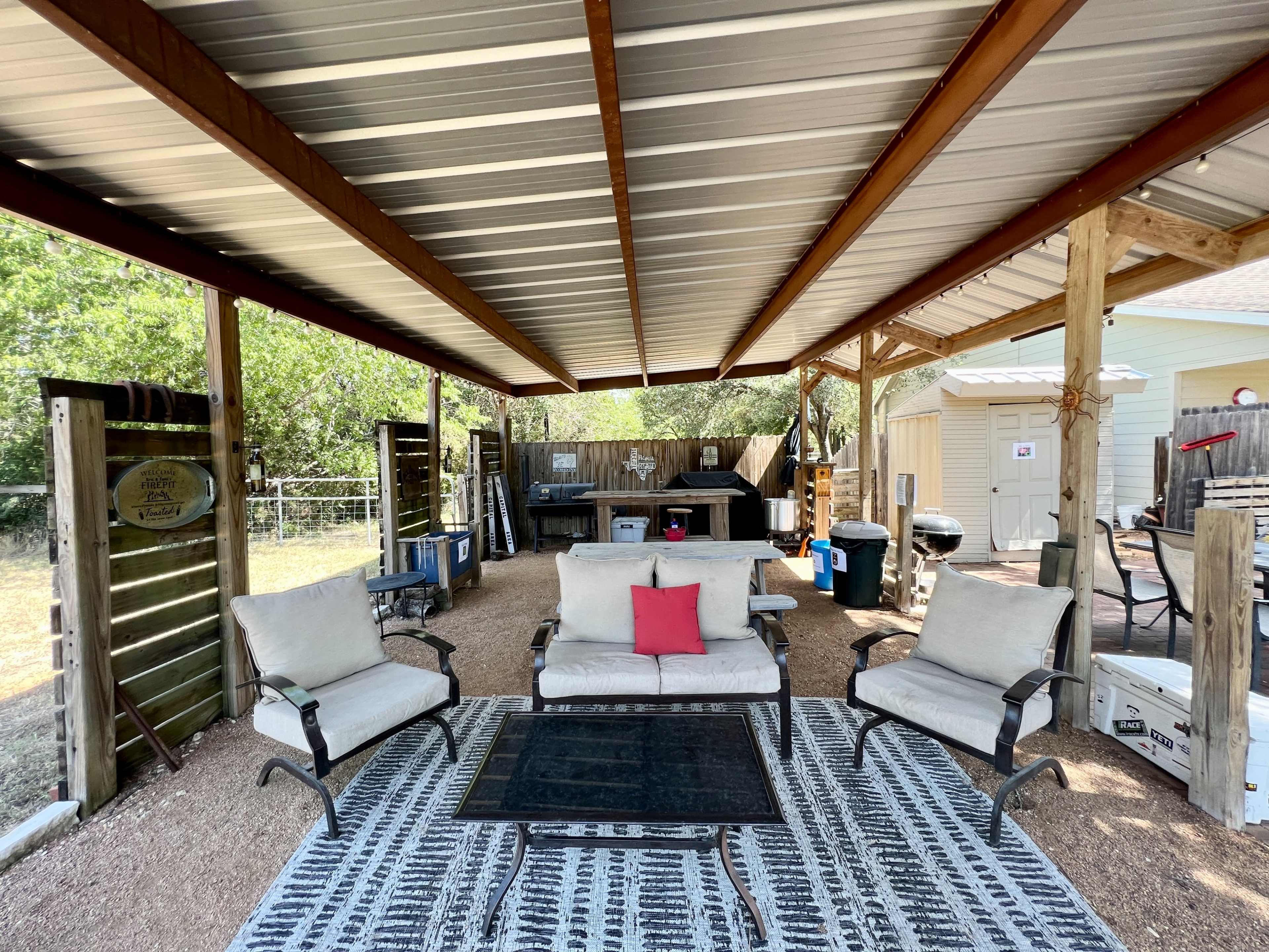 A covered outdoor seating area with two chairs and a table, surrounded by a gravel path and various outdoor storage items.