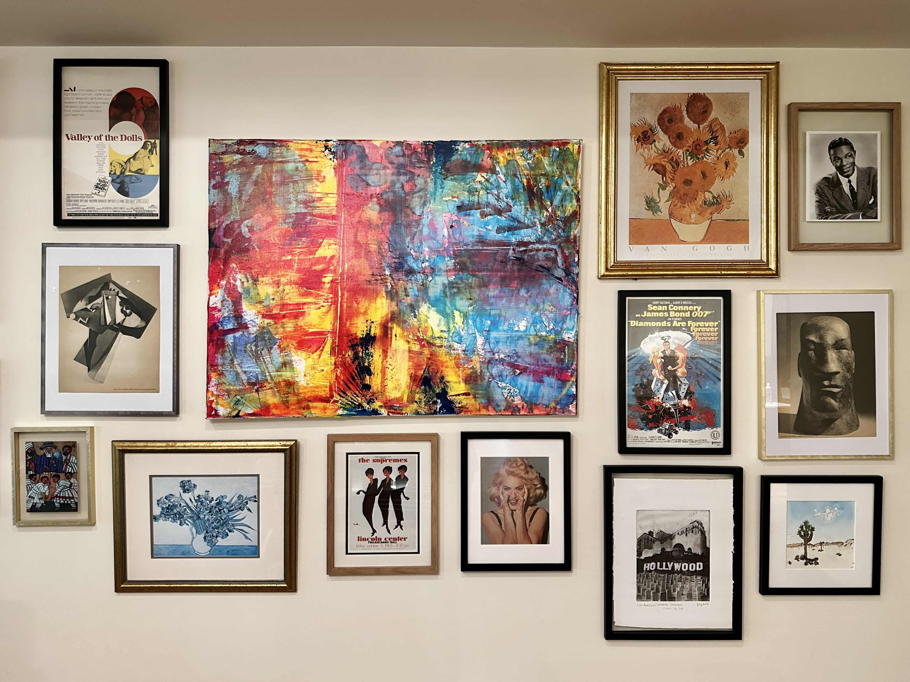 The image shows a decorative wall featuring a variety of framed artworks, including paintings, photographs, and posters in different styles and sizes.