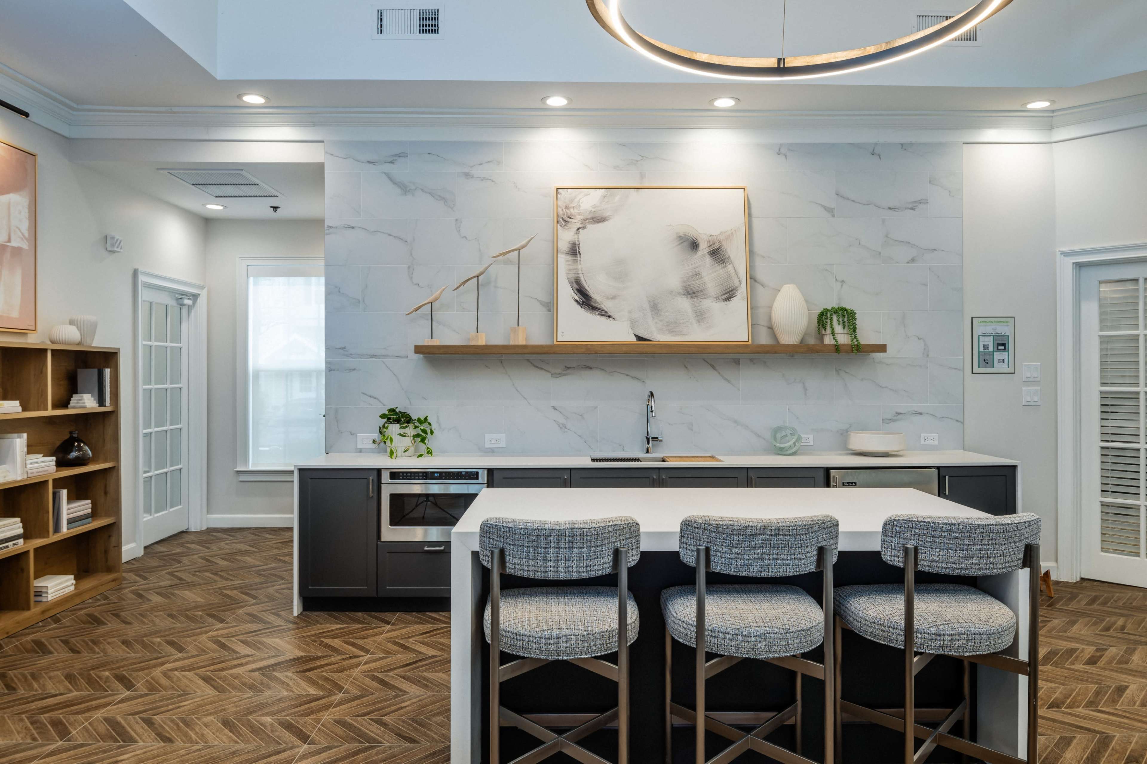 A modern kitchen with gray cabinetry, a white island bar with three upholstered stools, and a marble accent wall featuring a piece of artwork and a small shelf with a plant.