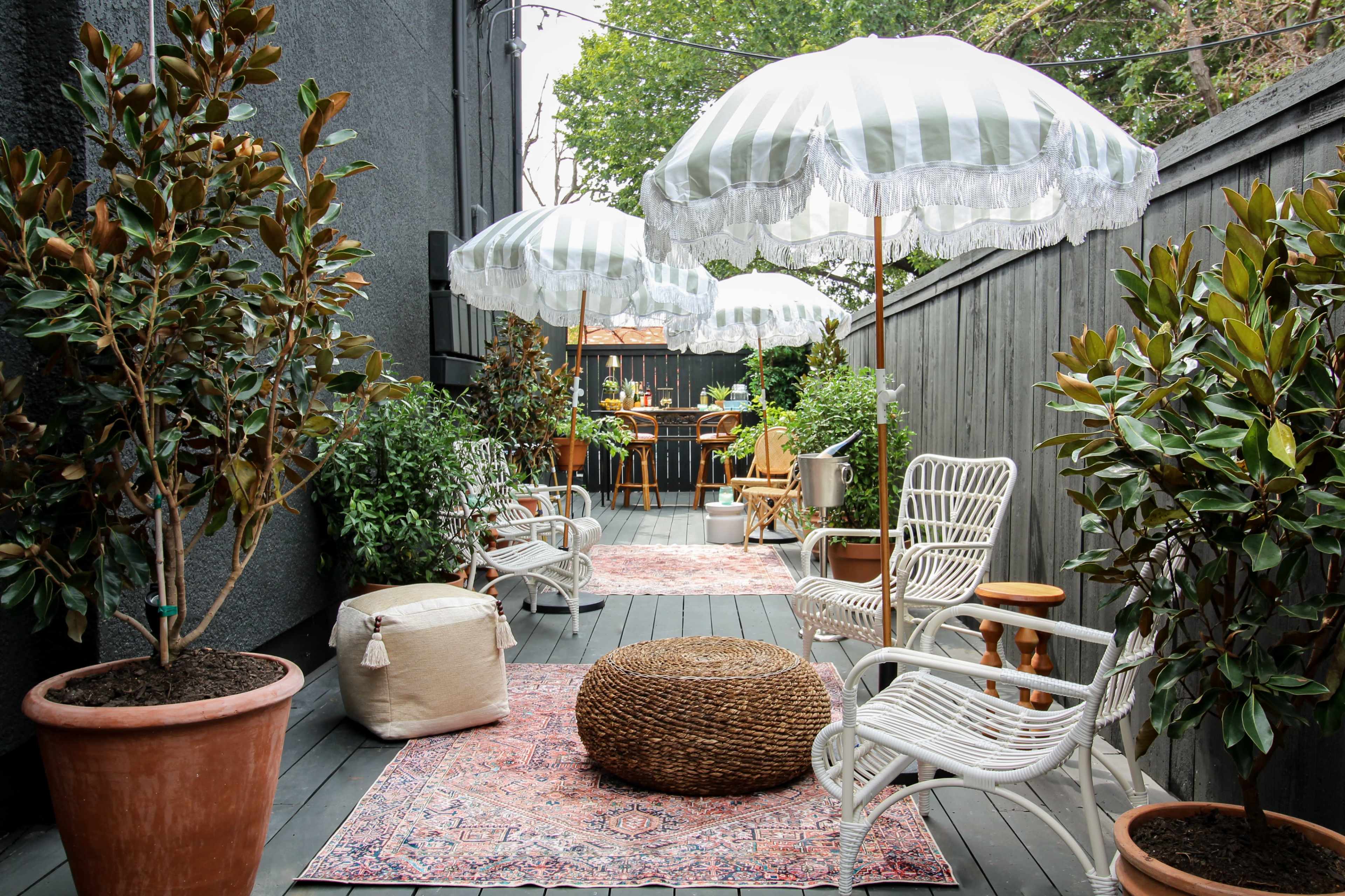 A narrow outdoor space featuring white wicker chairs, striped umbrellas, potted plants, and a patterned rug on wooden decking.