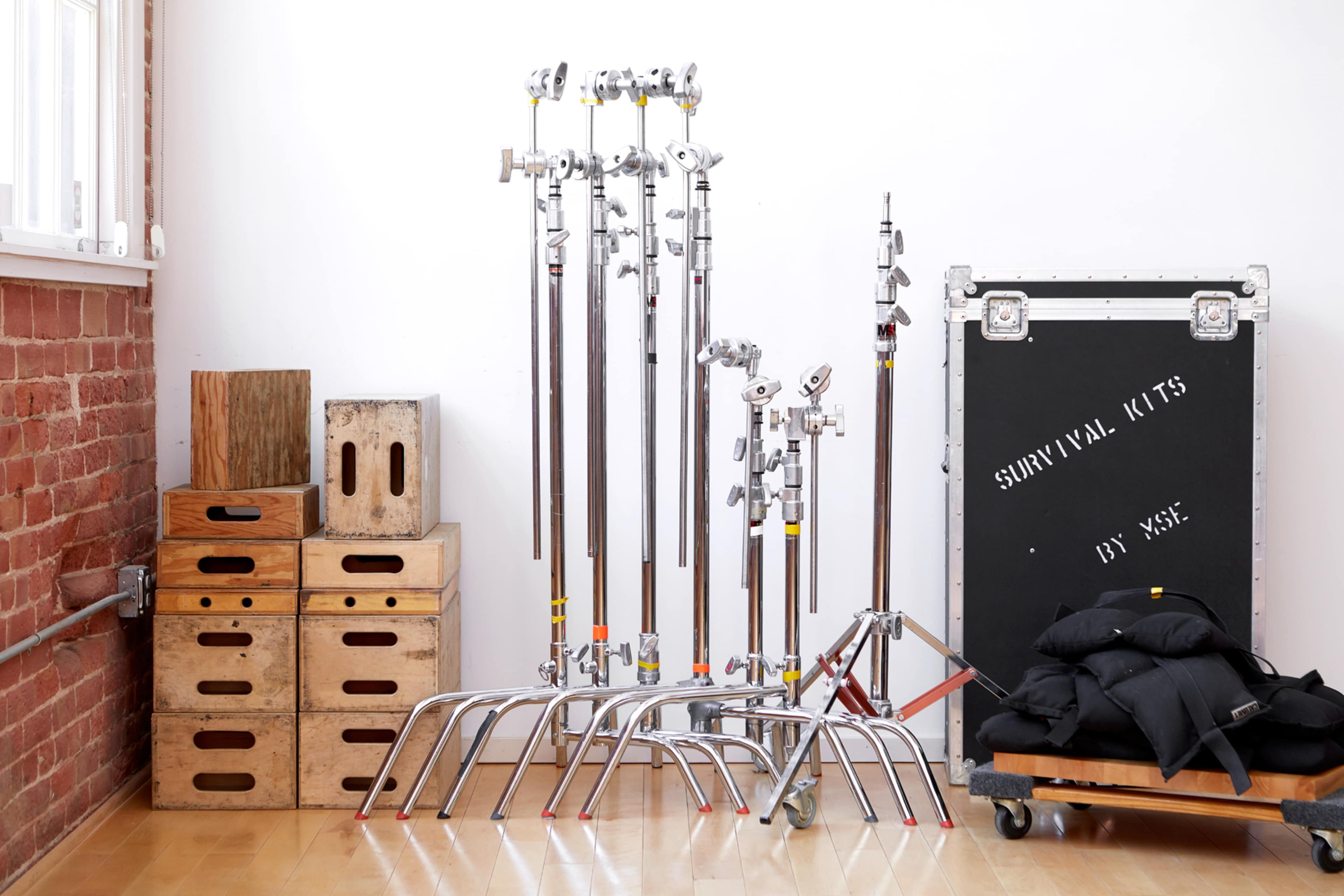 A collection of lighting stands, wooden crates, and a black equipment case labeled "SURVIVAL KITS" against a brick wall in a well-lit room.