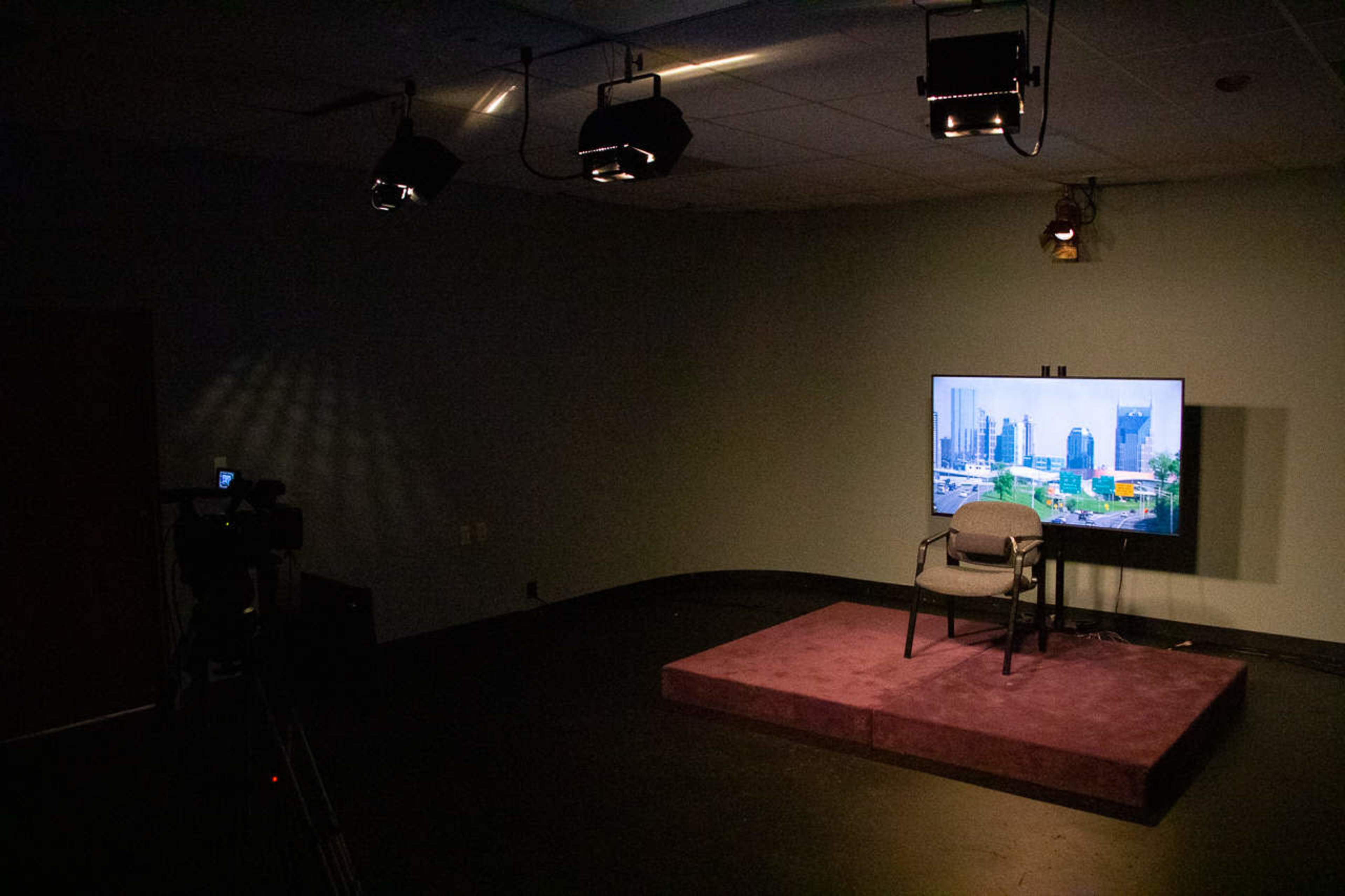 A dimly lit studio with a single chair on a purple stage, a television displaying a cityscape, and spotlights overhead.