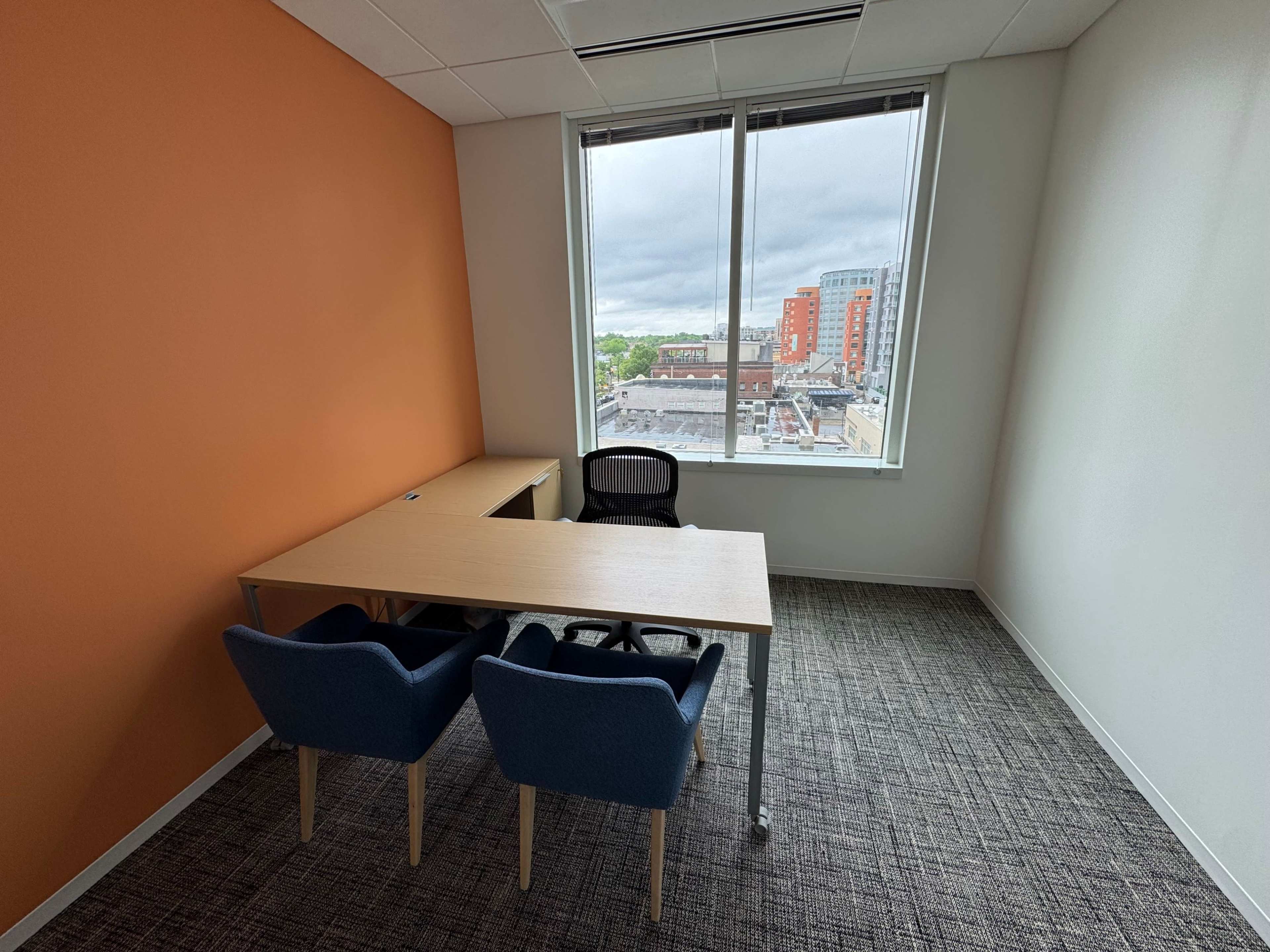 The image shows a small office space with an orange accent wall, a desk, two blue chairs, and large windows overlooking a cityscape.