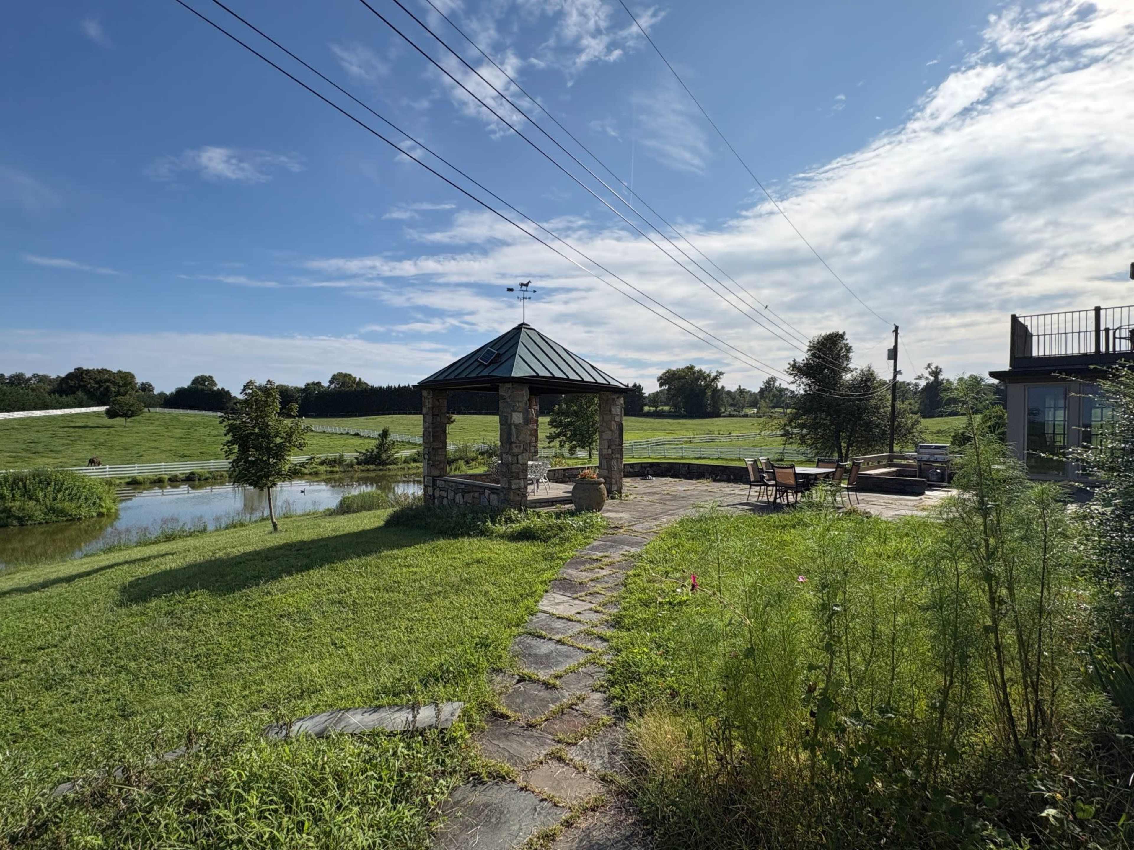 The image shows a gazebo near a pond surrounded by a grassy area, with a patio and outdoor furniture nearby, under a clear blue sky.