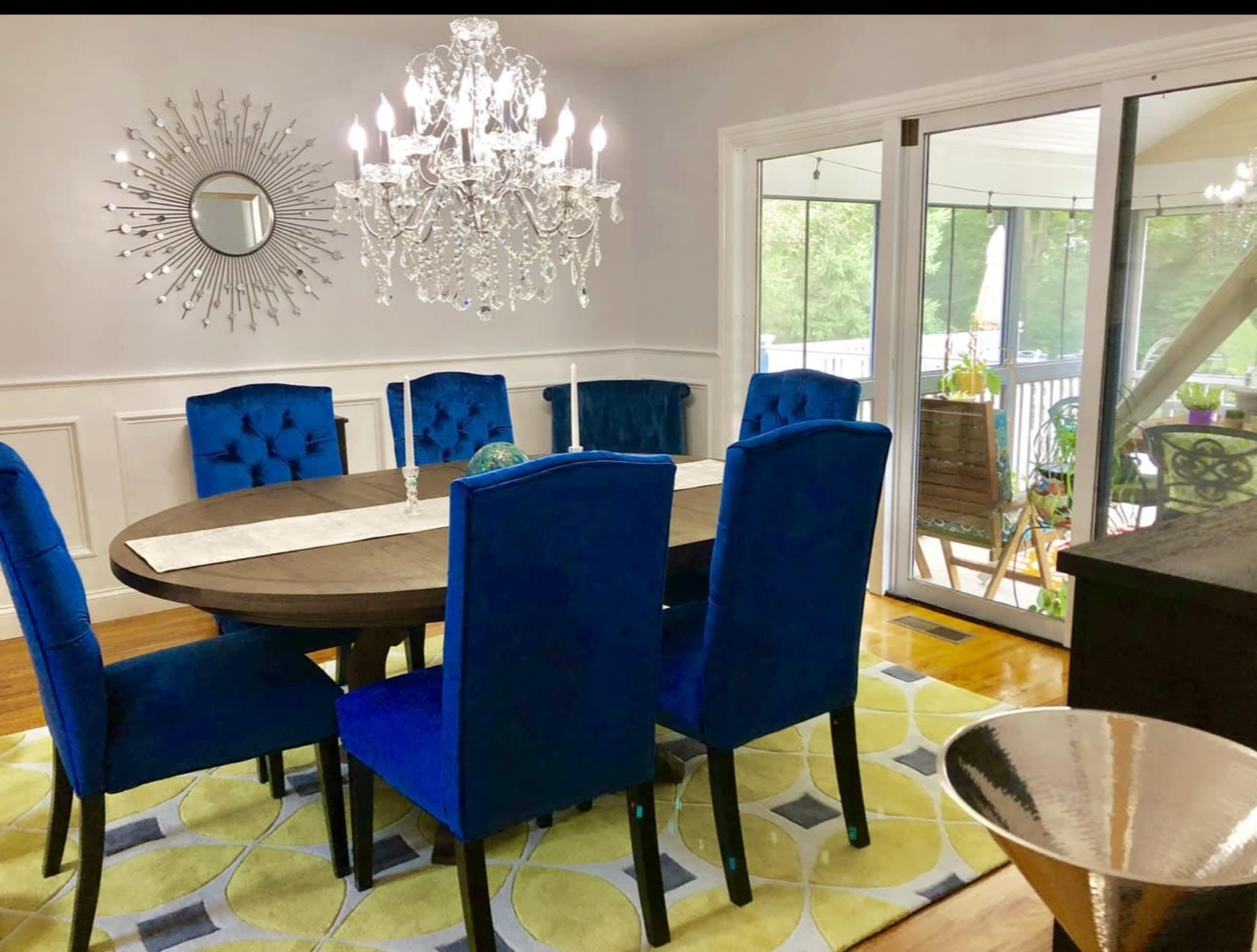 A dining room features a round table surrounded by blue upholstered chairs, under a decorative chandelier, with a large window opening to a deck area.